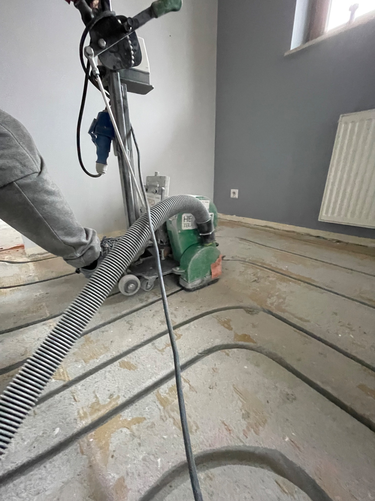 Szlifowanie podłoża pod ogrzewanie podłogowe maszyną z odkurzaczem. Widoczne rury instalacji w szlifowanej posadzce. Szare ściany i grzejnik w tle.
