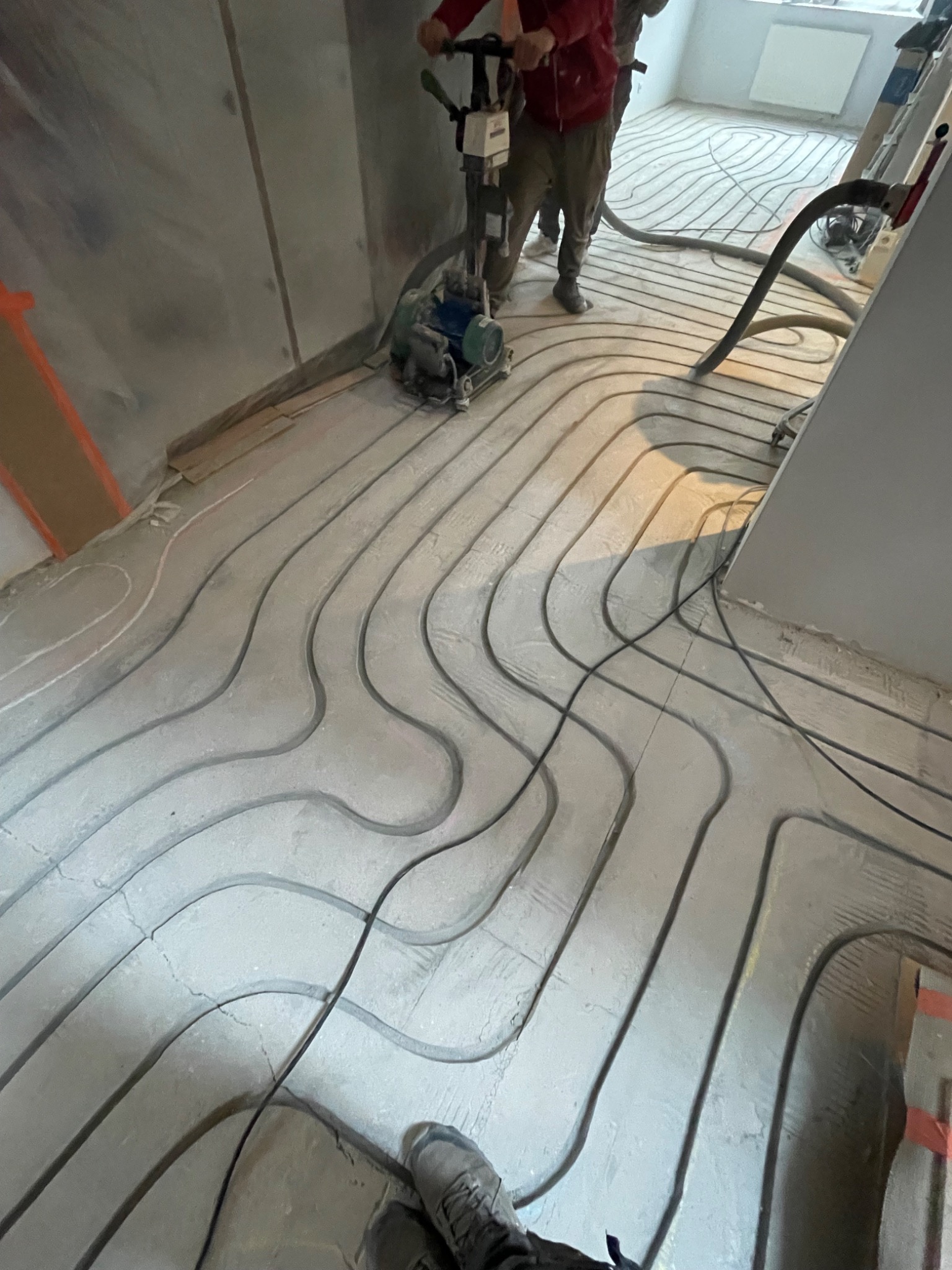 Układanie rur ogrzewania podłogowego w szarej wylewce, widoczne narzędzia i ekipa montażowa. Rury ułożone w pętle, przygotowane do zalania posadzką.