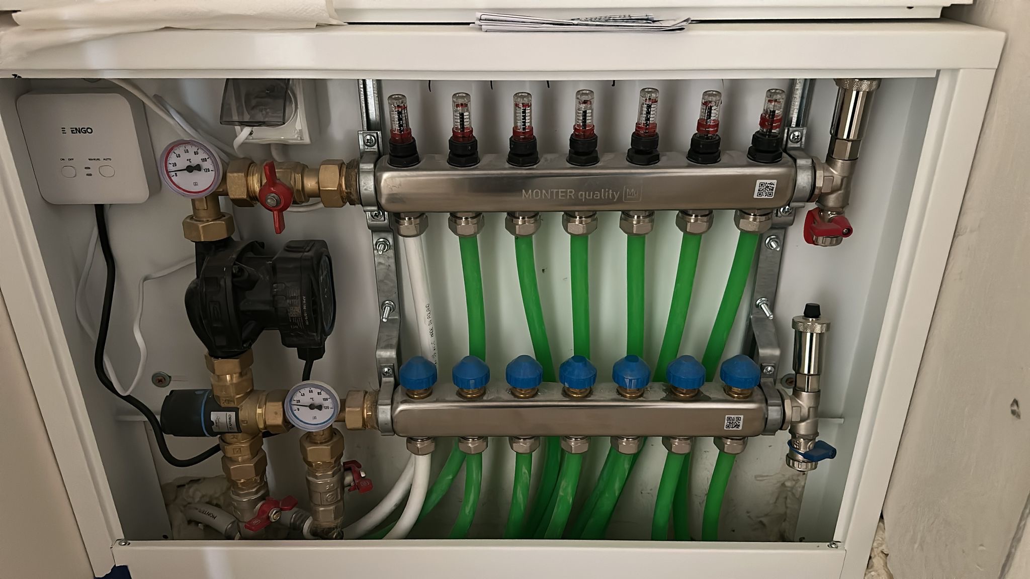 Rozdzielacz ogrzewania podłogowego w szafce, z zielonymi rurami i elementami sterującymi. Widoczne manometry, pompa obiegowa i zawory. Górny rozdzielacz z przepływomierzami.