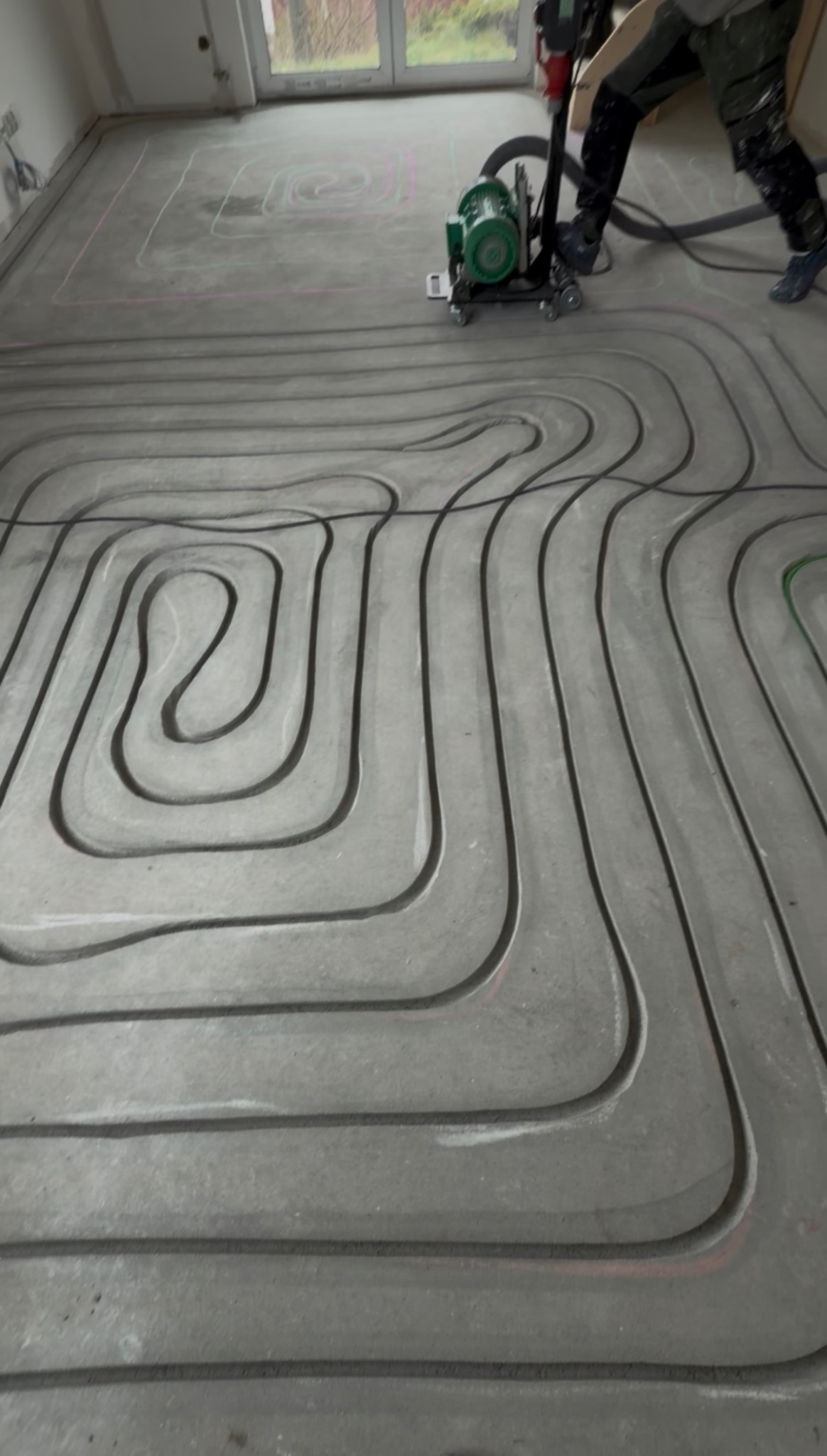 Frezowanie bruzd w posadzce pod ogrzewanie podłogowe. Widoczne równe, wyfrezowane kanały i fragment frezarki. Posadzka przygotowana do montażu rur grzewczych.