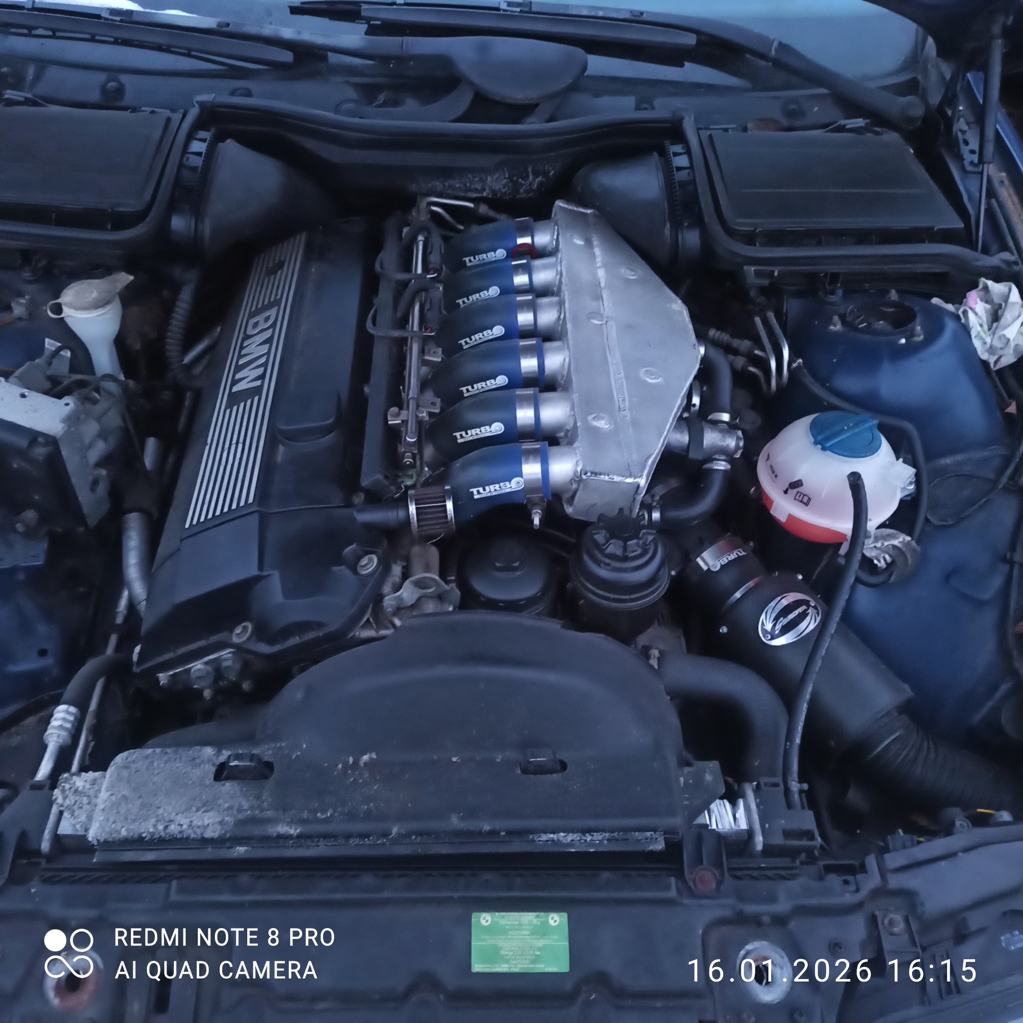 Silnik BMW po tuningu z widocznymi turbosprężarkami, srebrną kolektorem dolotowym i niebieskimi przewodami. Widok z góry na otwartą maskę samochodu.