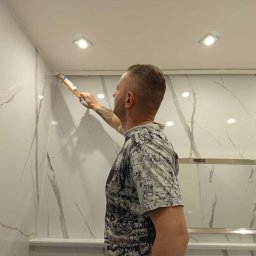 Zajdex - Mężczyzna aplikuje silikon na styku ściany z marmurowym wzorem i sufitu w łazience, używając pistoletu do silikonu. Widoczne oświetlenie sufitowe.