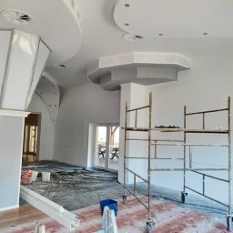 Zajdex - Wnętrze w trakcie malowania: białe ściany, dekoracyjne elementy sufitowe, rusztowanie malarskie, zabezpieczona podłoga. Widoczne narzędzia i materiały malarskie.