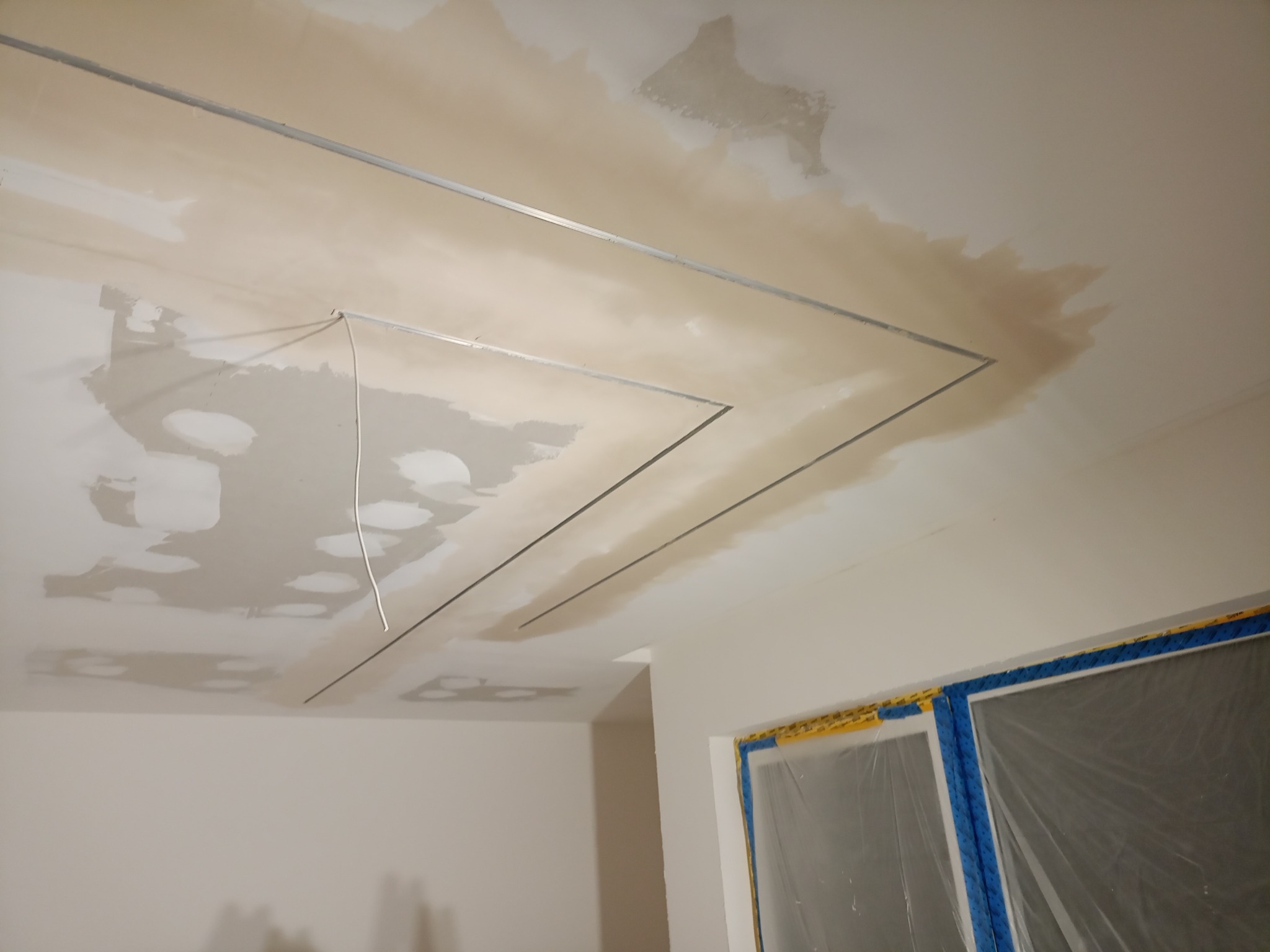 Sufit z metalową konstrukcją pod płyty gipsowe i częściowo nałożoną gładzią, okno zabezpieczone folią malarską. Etap prac wykończeniowych.