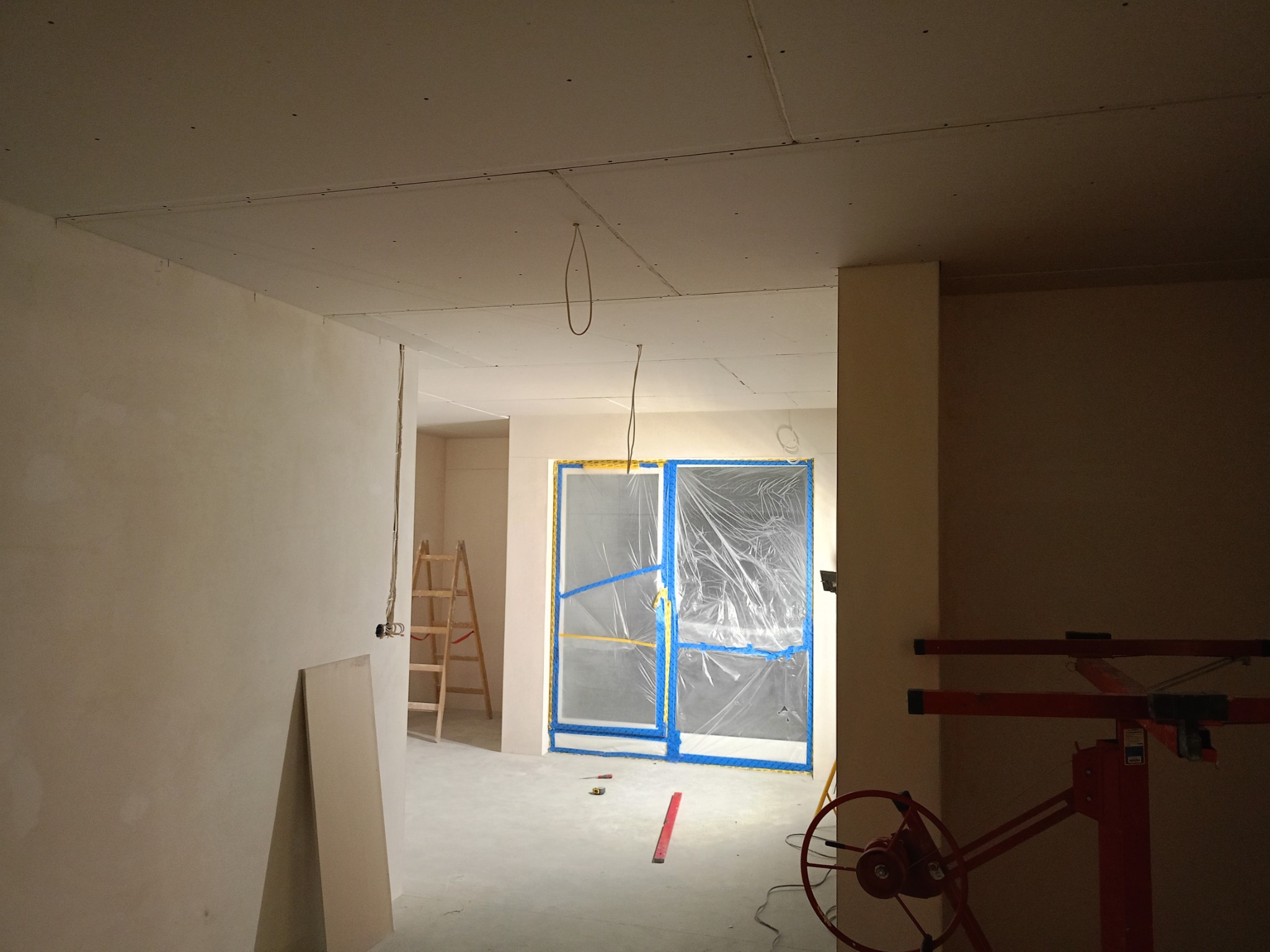 Wnętrze w trakcie prac wykończeniowych: zamontowana płyta gips-karton na suficie, zabezpieczone folią drzwi, drabina i podnośnik do płyt widoczne w pomieszczeniu.