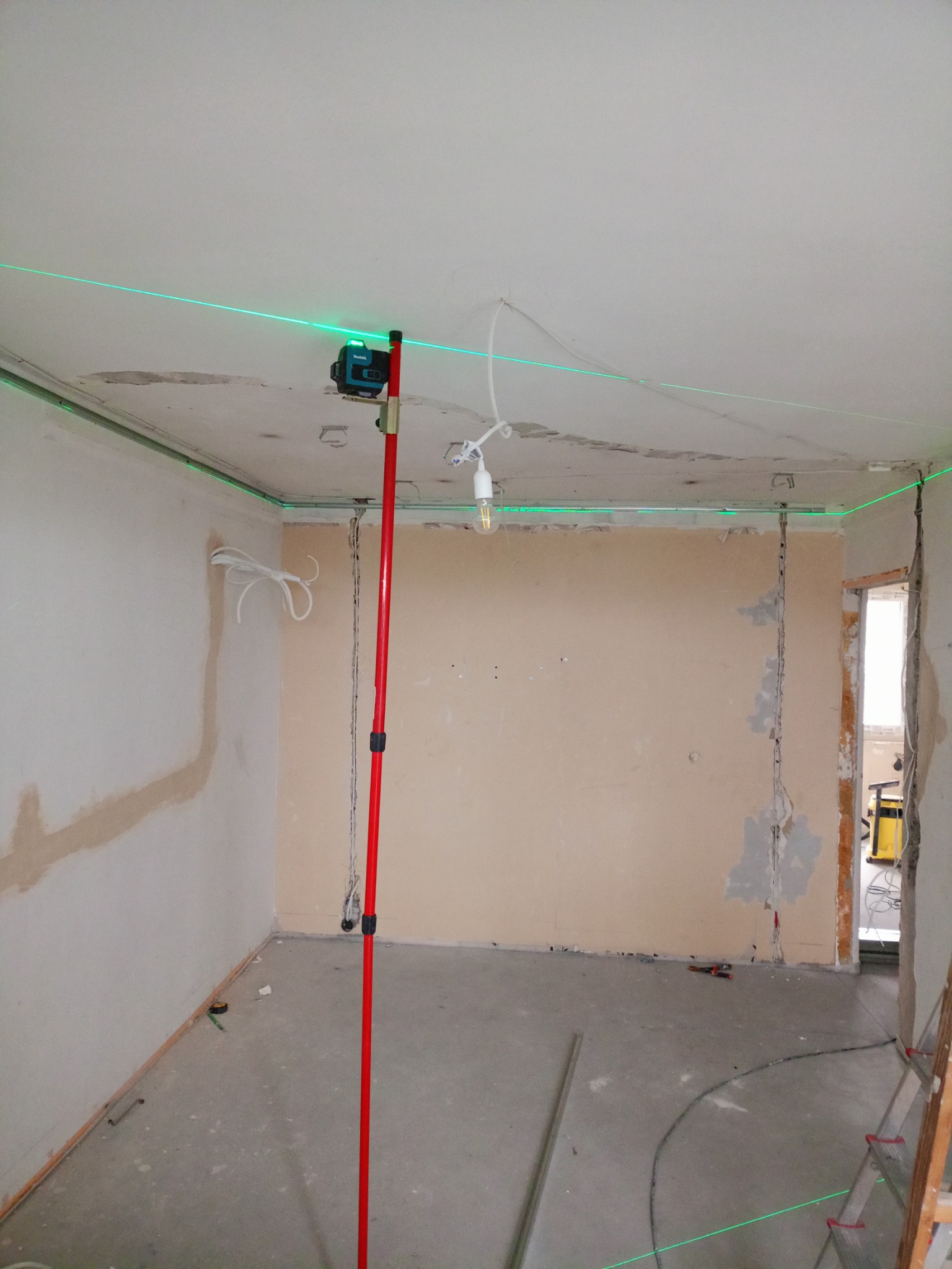 Pokój w trakcie remontu: ściany przygotowane do gładzi, widoczny laser krzyżowy wyznaczający poziom, surowy beton na podłodze. Prace wykończeniowe w toku.