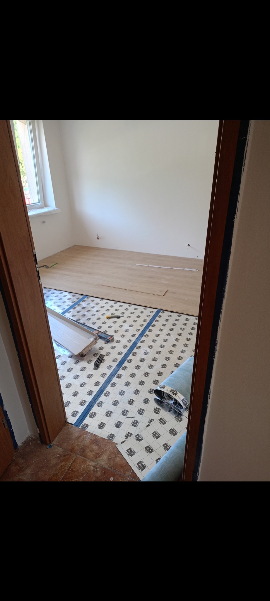 Układanie paneli podłogowych w pokoju, widok zza futryny. Część podłogi pokryta podkładem, panele częściowo ułożone. Widoczne narzędzia i resztki materiałów.