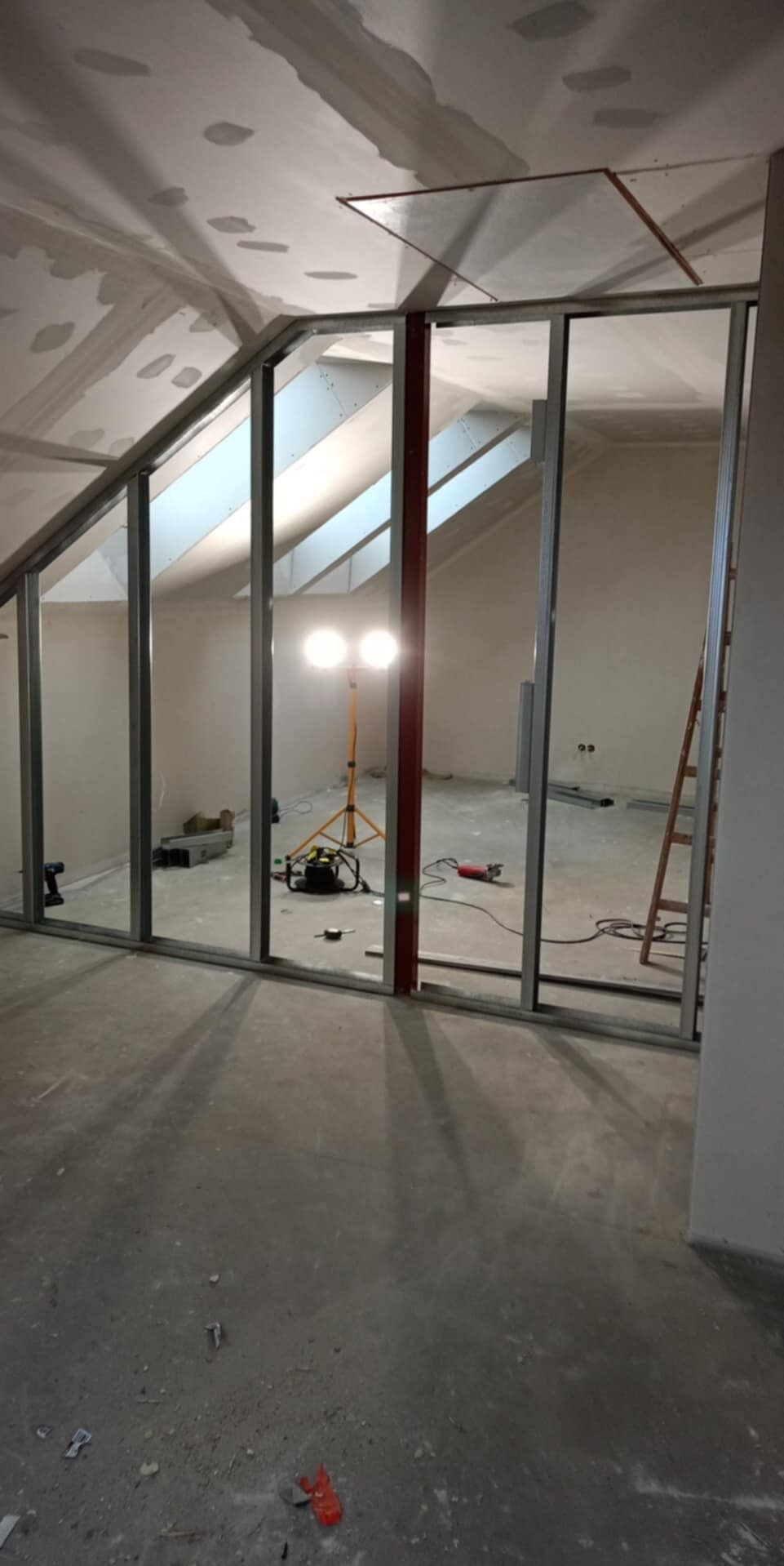 Stelaż z profili metalowych pod ściankę działową z płyty karton-gips na poddaszu. Widoczne narzędzia i lampa budowlana. Skos dachu z oknami.