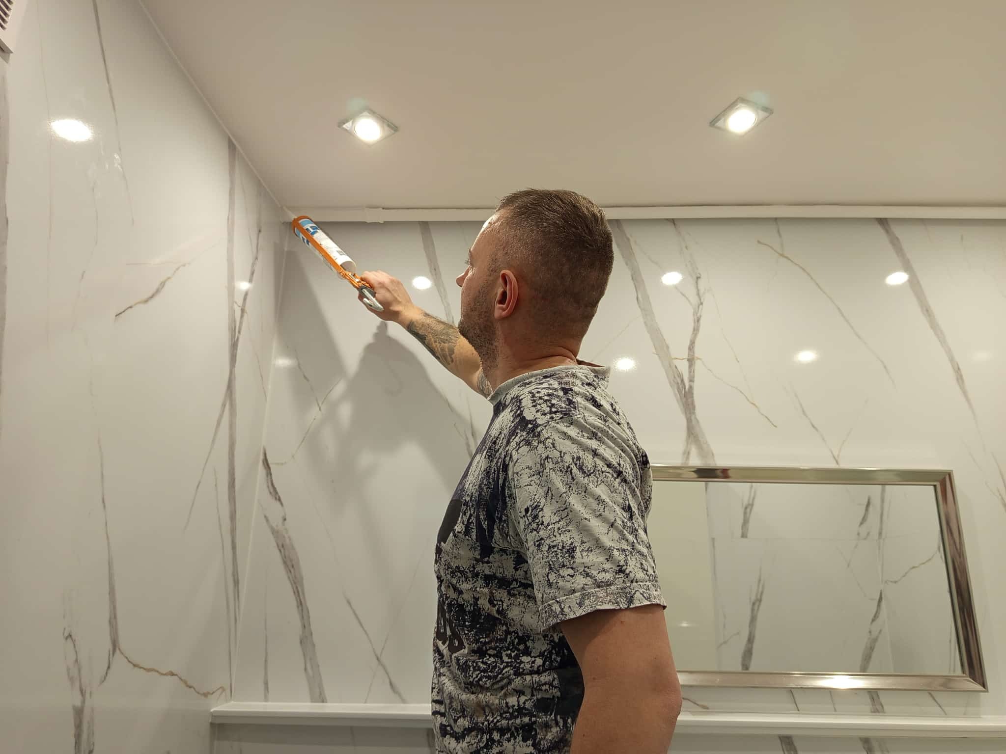 Mężczyzna aplikuje silikon na styku ściany z marmurowym wzorem i sufitu w łazience, używając pistoletu do silikonu. Widoczne oświetlenie sufitowe.