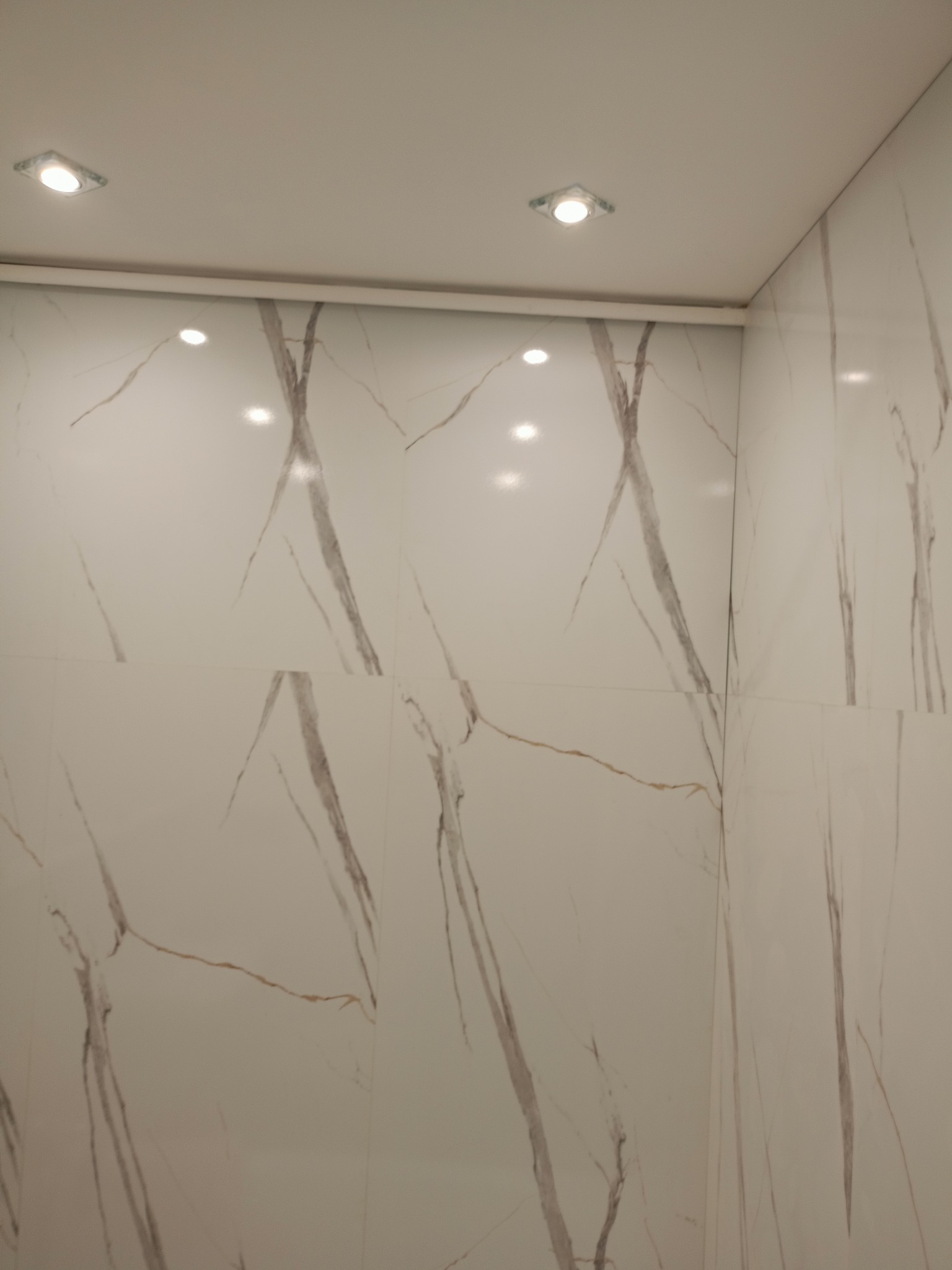 Białe, błyszczące płytki imitujące marmur z szarymi żyłami na ścianie i suficie, oświetlone punktowymi lampami LED. Wykończenie wnętrza łazienki.