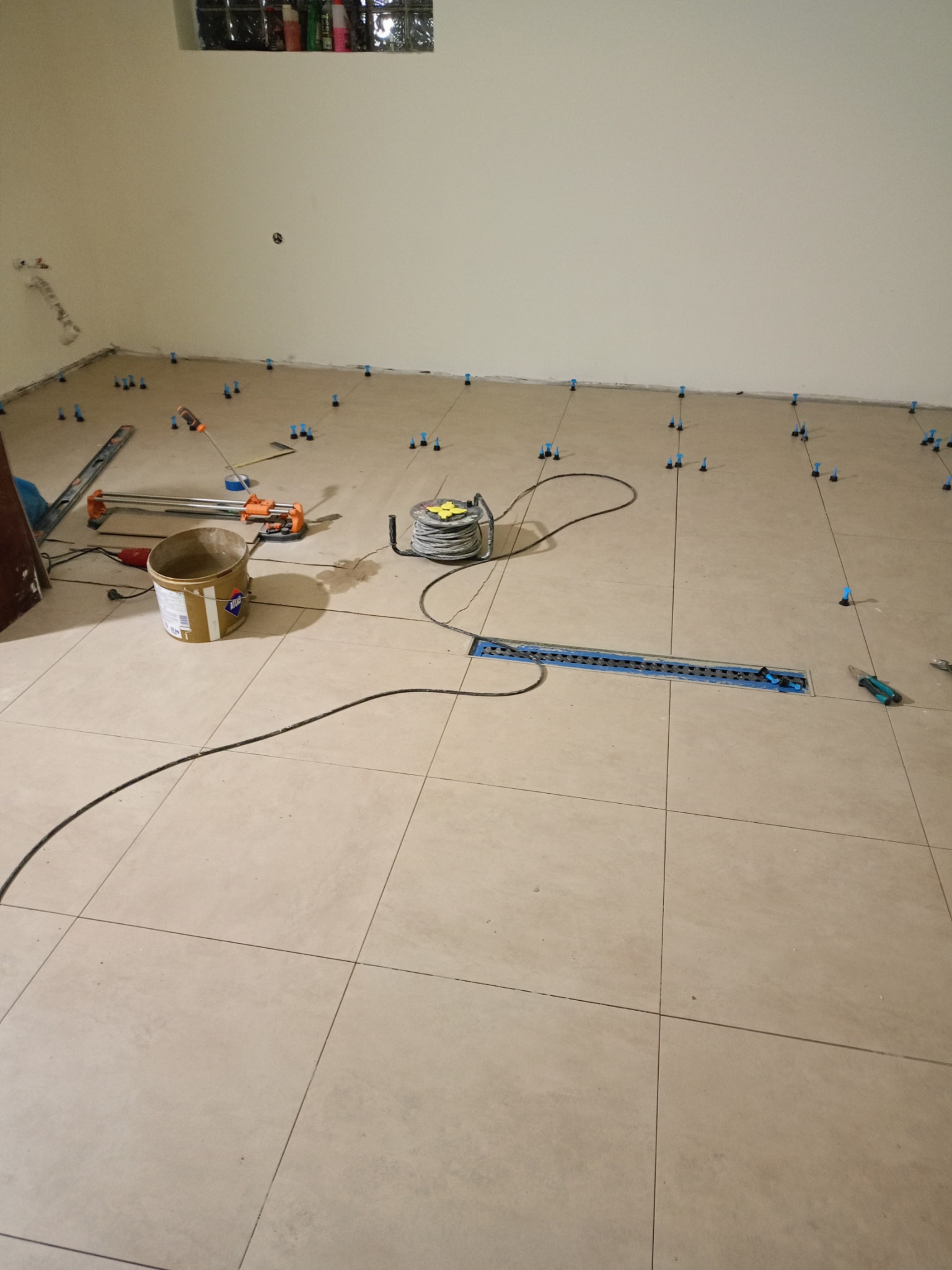 Układanie beżowych płytek podłogowych z użyciem systemu poziomowania. Widoczne narzędzia: wiadro, poziomica, przecinarka do płytek. Etap prac przed fugowaniem.