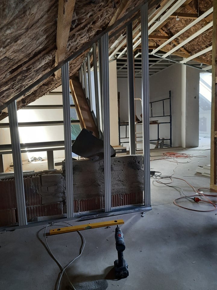 Montaż metalowej konstrukcji ścianki działowej na poddaszu. Widoczne narzędzia: poziomica i wkrętarka. Ceglana ściana w tle. Ocieplenie poddasza wełną mineralną.