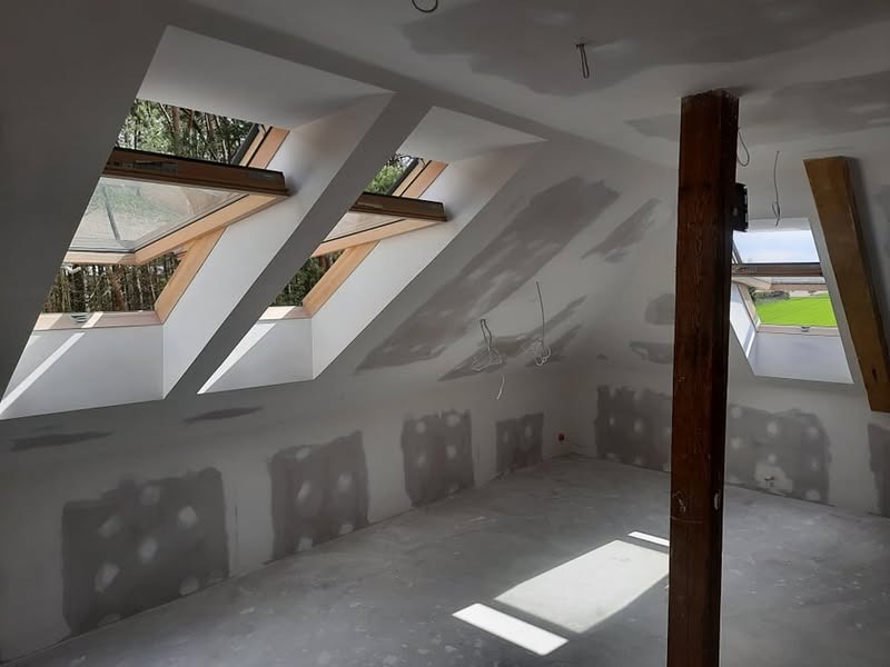 Poddasze w trakcie prac wykończeniowych: szpachlowane ściany, drewniany słup konstrukcyjny, okna dachowe z widokiem na zieleń. Wnętrze do aranżacji.