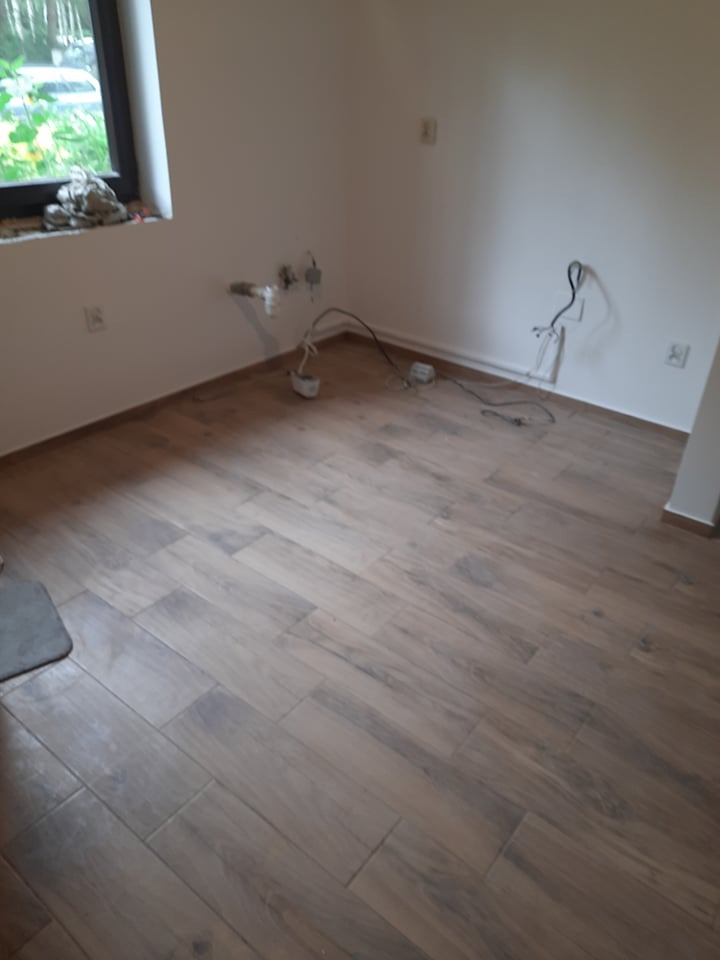 Ukończona podłoga z płytek imitujących drewno w pustym pokoju z instalacjami na ścianach. Widok na okno z zielenią na zewnątrz. Minimalistyczne wnętrze po remoncie.