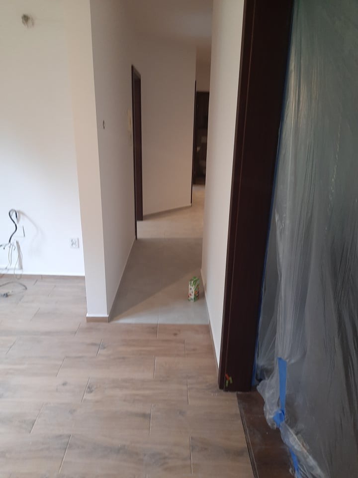 Korytarz po remoncie z drewnopodobną podłogą, świeżo pomalowanymi ścianami i zabezpieczonymi folią drzwiami. Widoczne kable i karton soku.