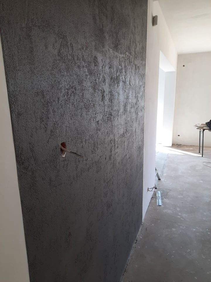 Ściana z dekoracyjną, szarą strukturą, widoczne gniazdko elektryczne z wystającymi przewodami. W tle jasne pomieszczenie w trakcie wykończenia.