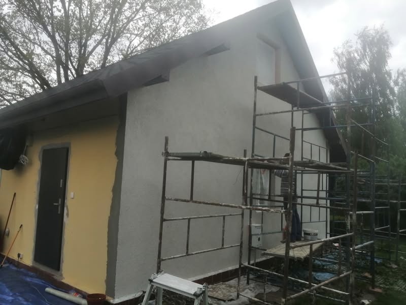 Elewacja domu w trakcie prac: część ściany otynkowana na szaro, druga w kolorze żółtym. Widoczne rusztowania i narzędzia malarskie. Prace wykończeniowe.