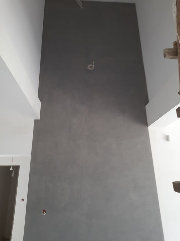 Szara ściana z efektem dekoracyjnym w surowym wnętrzu, widoczne kable i fragmenty konstrukcji, świeżo po malowaniu, z delikatną fakturą i minimalistycznym wykończeniem.