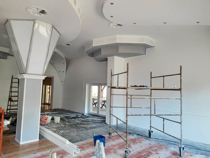 Wnętrze w trakcie malowania: białe ściany, dekoracyjne elementy sufitowe, rusztowanie malarskie, zabezpieczona podłoga. Widoczne narzędzia i materiały malarskie.