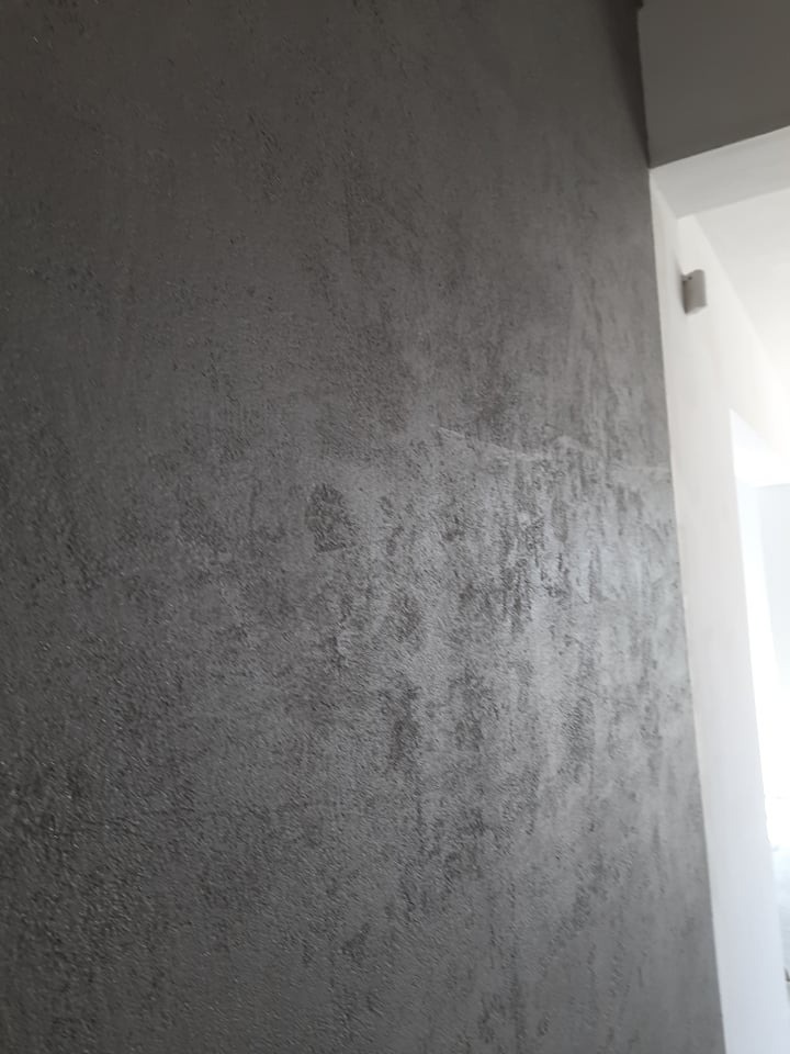 Szara ściana z efektem dekoracyjnym, imitującym beton, w nowoczesnym wnętrzu. Widoczny fragment białego narożnika i sufitu.