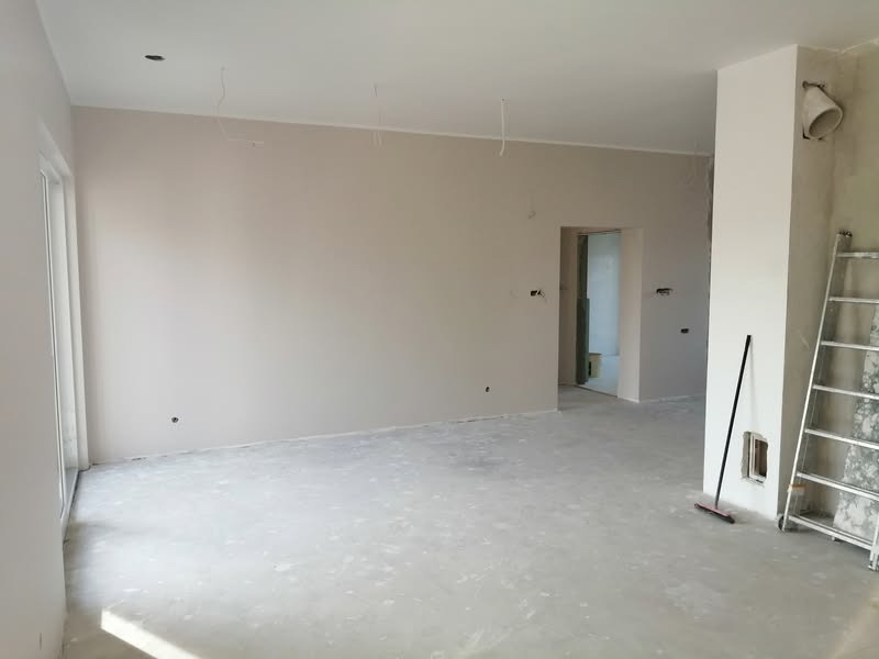 Jasne, przestronne wnętrze w trakcie wykańczania: świeżo pomalowane ściany w neutralnym odcieniu, surowa podłoga, drabina oparta o filar. Widoczne kable i otwory.