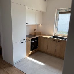 WYMIAROWE MEBLE SEBASTIAN HABERSKI - Nowoczesna kuchnia z białymi szafkami górnymi i wysokimi, oraz drewnianymi szafkami dolnymi. Minimalistyczne uchwyty, płyta indukcyjna i piekarnik w zabudowie.