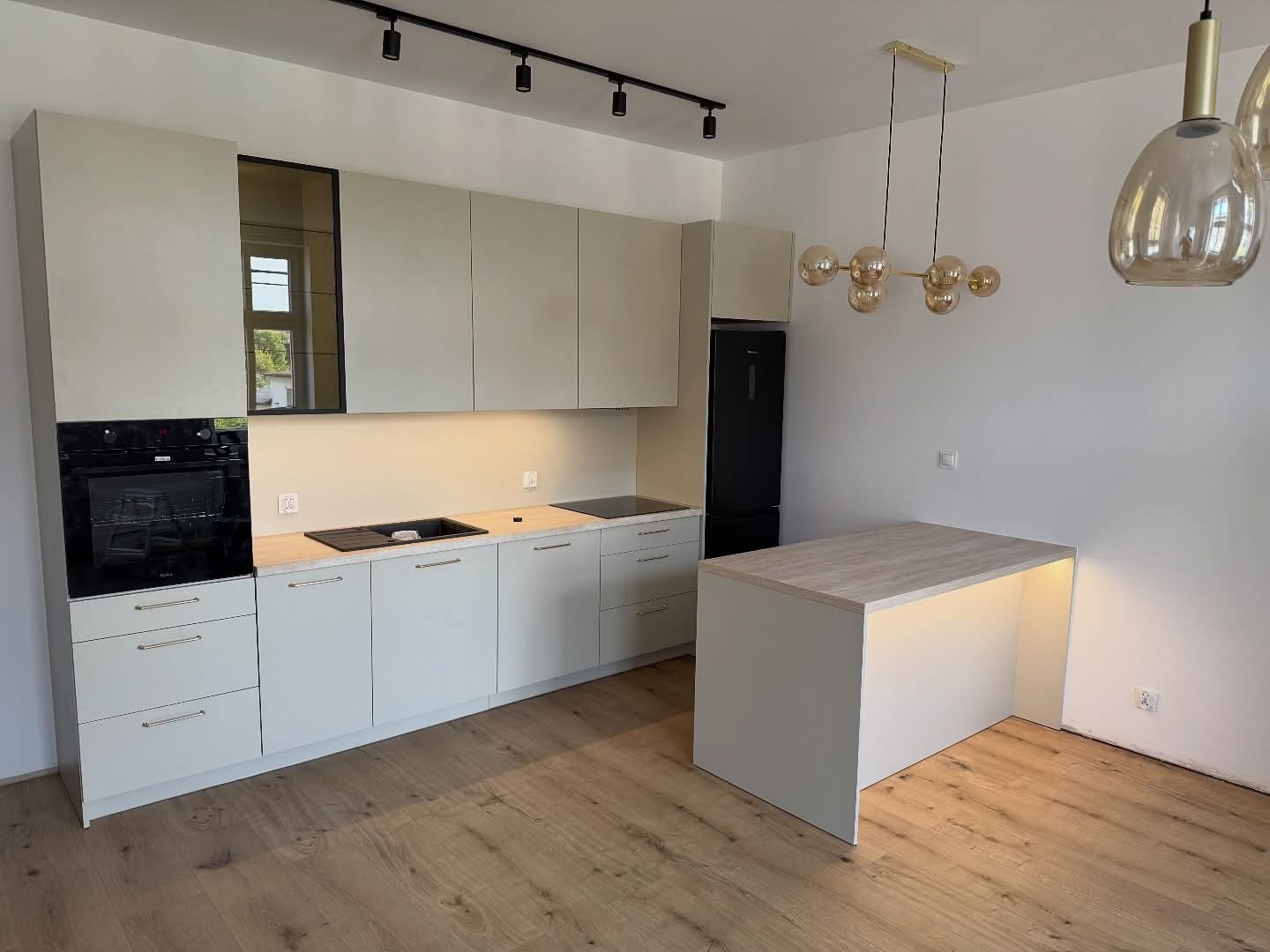 Jasna kuchnia z wyspą, fronty bez uchwytów, czarny piekarnik i lodówka. Oświetlenie podszafkowe i lampy wiszące. Parkiet w jodełkę. Minimalistyczny design.