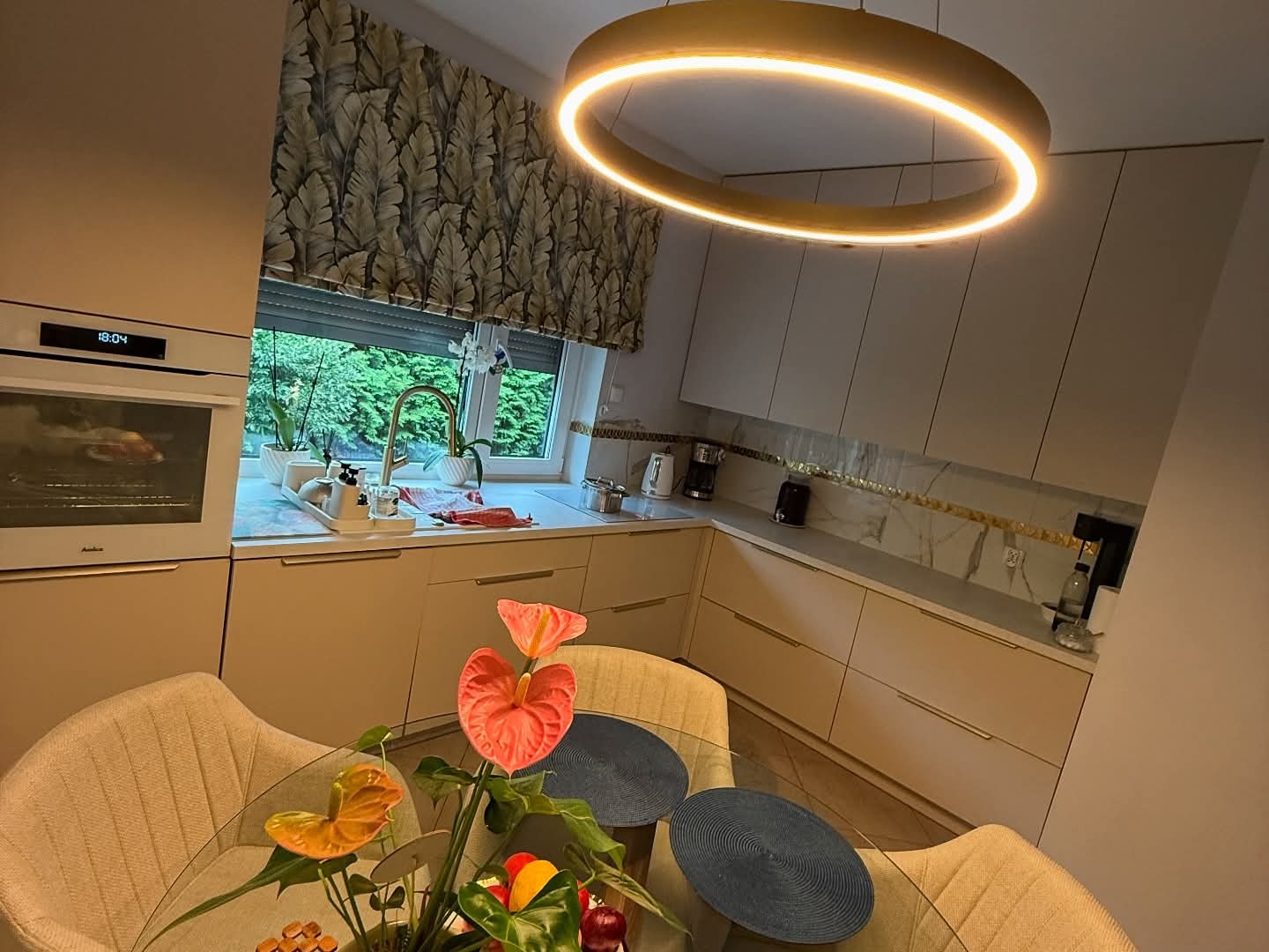 Jasna kuchnia z beżowymi frontami, marmurowym blatem i okrągłą lampą. Na pierwszym planie kwiaty w wazonie na szklanym stole. Widok na okno z zielenią.