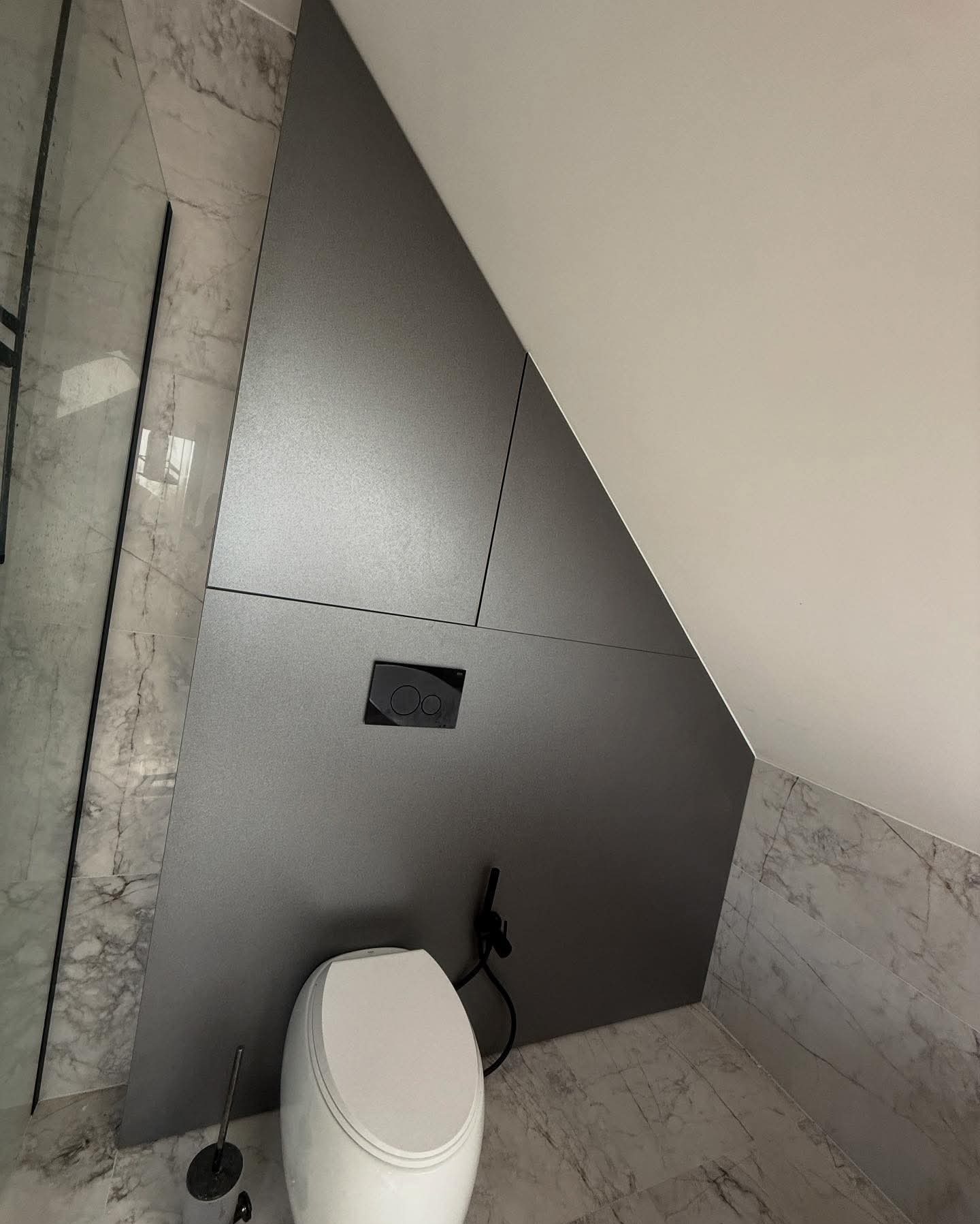 Szara zabudowa wnęki w łazience ze spłuczką podtynkową i toaletą. Ukośna ściana, marmurowe płytki. Nowoczesny design.