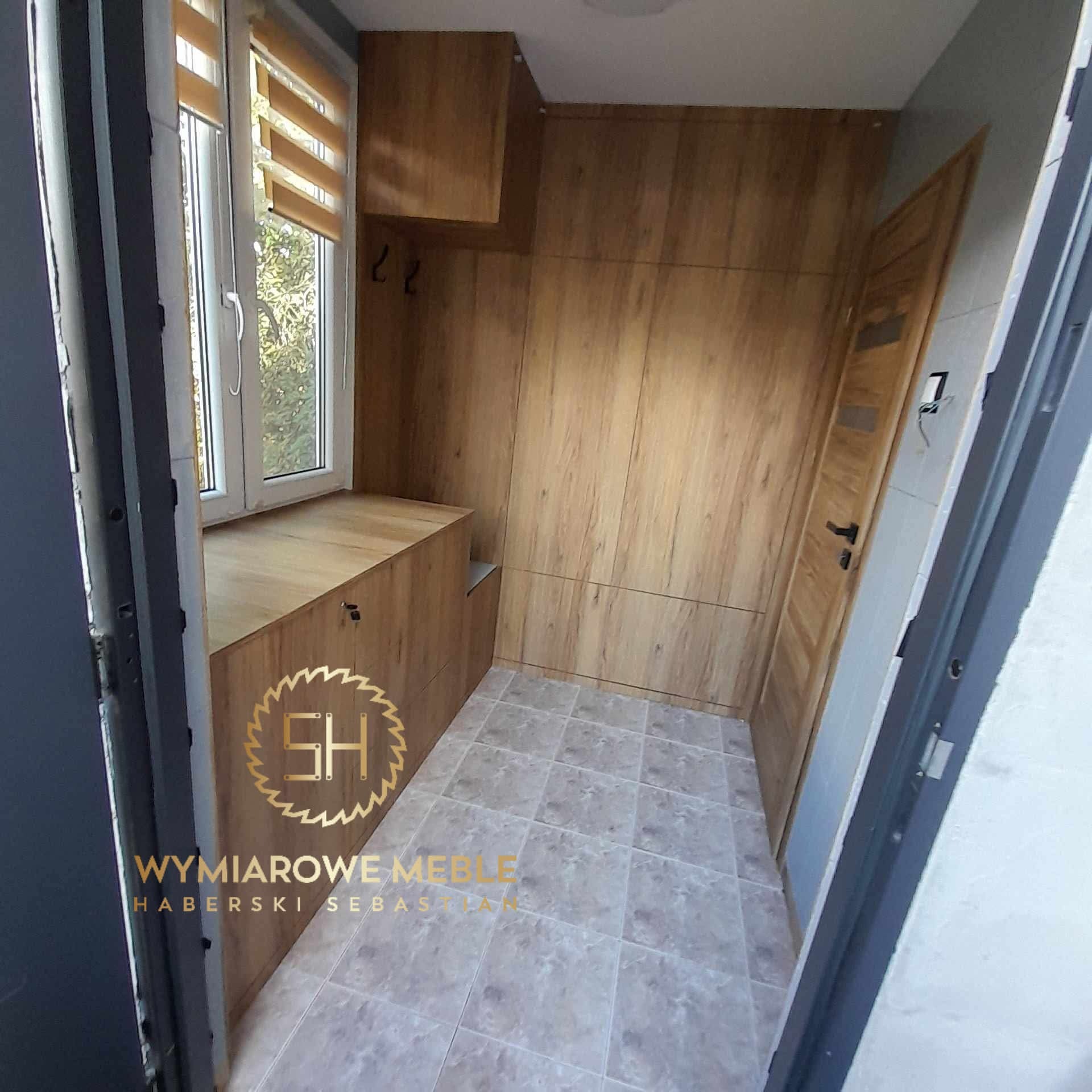 Wnęka z zabudową z drewna w odcieniach brązu, z szafkami pod oknem i szafą wzdłuż ściany. Widoczna drewniana podłoga i drzwi. Ujęcie z perspektywy wejścia.