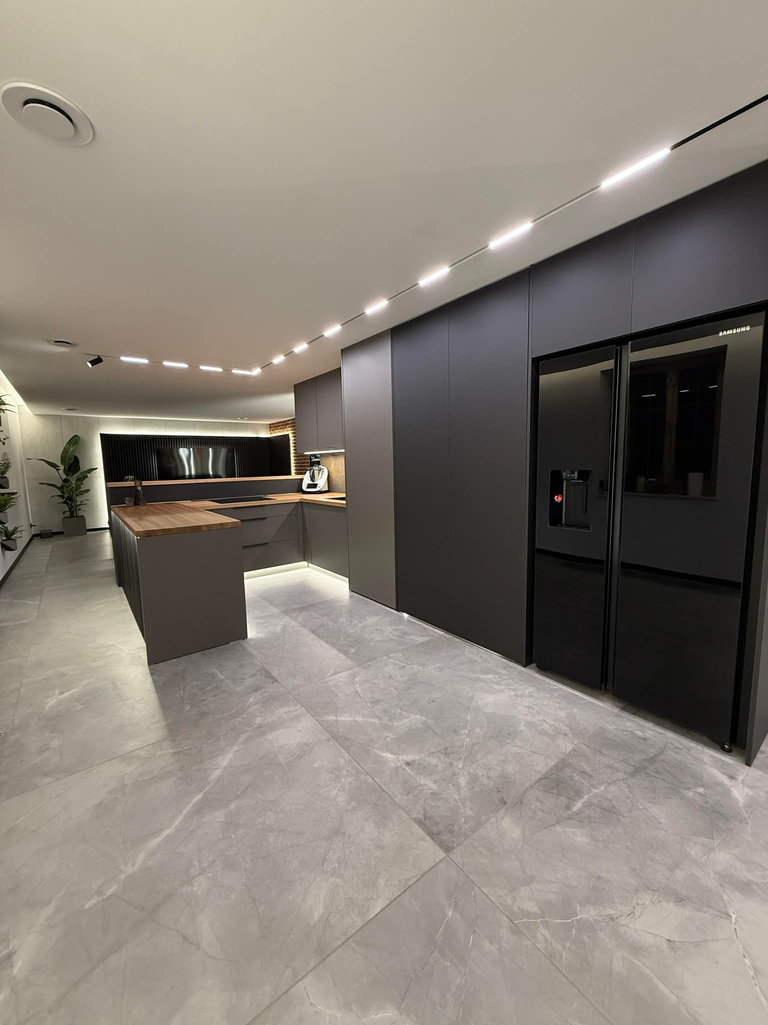 Nowoczesna kuchnia w odcieniach szarości z drewnianym blatem wyspy, oświetleniem LED i dużą lodówką Samsung side-by-side. Minimalistyczny design i marmurowe płytki podłogowe.