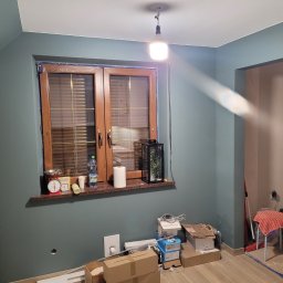 Piotr Zjeżdżałka Układanie Glazury i Terakoty - Pokój w trakcie malowania ścian na kolor morski, widoczne okno z drewnianą ramą, kartony na podłodze i prowizoryczne oświetlenie. Ściana w kolorze morskim.