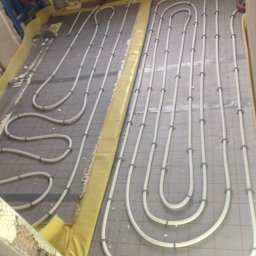 Piotr Zjeżdżałka Układanie Glazury i Terakoty - Białe rury ogrzewania podłogowego, zamocowane na szarej macie z siatką, układane w pętle. Żółta folia izolacyjna przy ścianach. Widoczne fragmenty instalacji hydraulicznej.