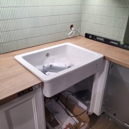 Piotr Zjeżdżałka Kompleksowe Remonty, Wykończenia i Hydraulika, ponad 35 lat doświadczenia - Biała, ceramiczna umywalka wpuszczana w drewniany blat, z widoczną instalacją elektryczną i poziomnicą, na tle ściany wyłożonej podłużnymi, zielonymi płytkami.