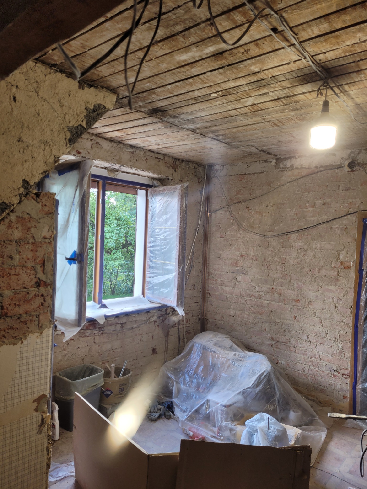 Pokój w trakcie remontu: odsłonięta cegła, zabezpieczone okno folią, widoczne instalacje, surowy strop. Przygotowania do dalszych prac wykończeniowych.