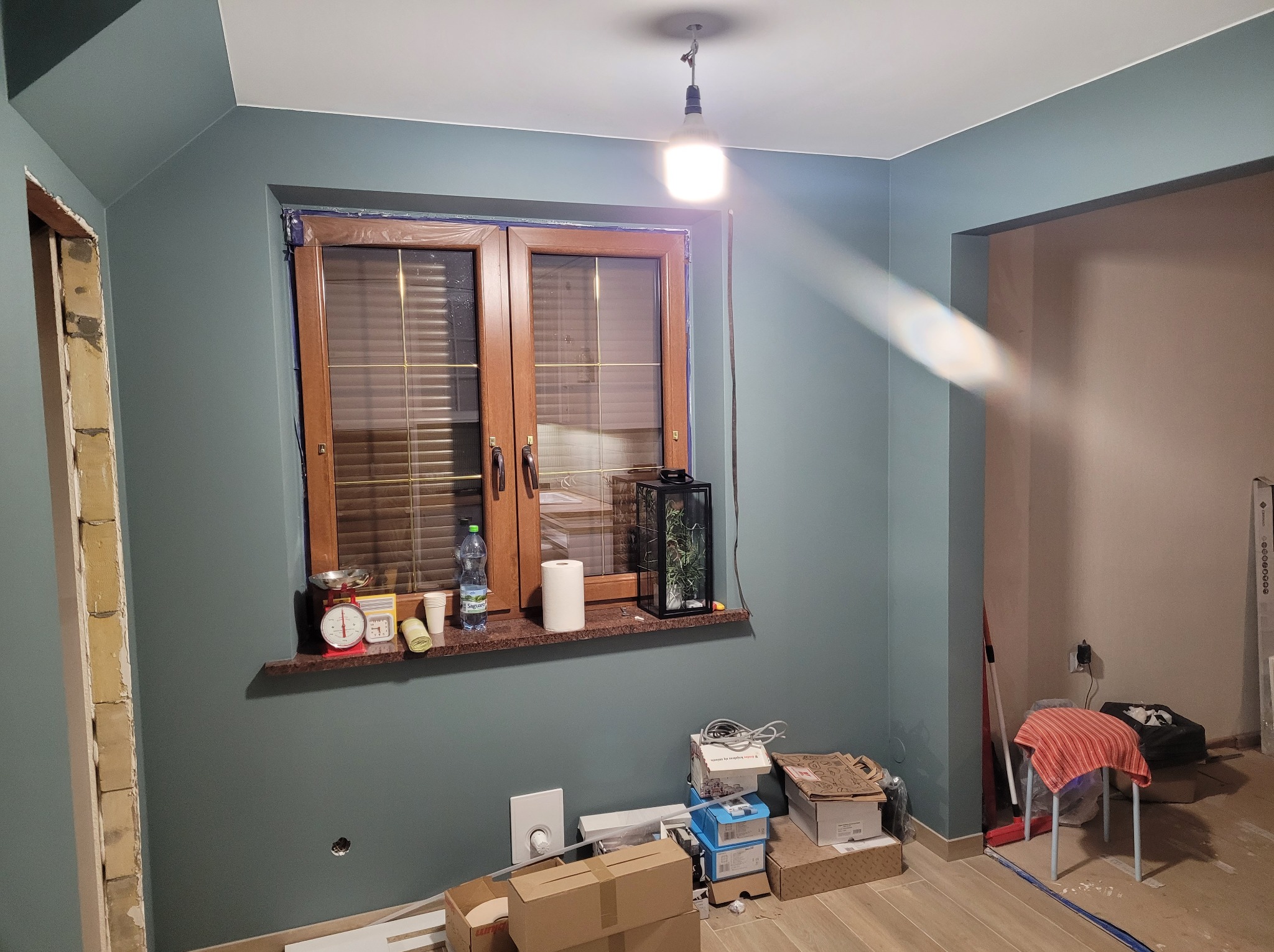 Pokój w trakcie malowania ścian na kolor morski, widoczne okno z drewnianą ramą, kartony na podłodze i prowizoryczne oświetlenie. Ściana w kolorze morskim.