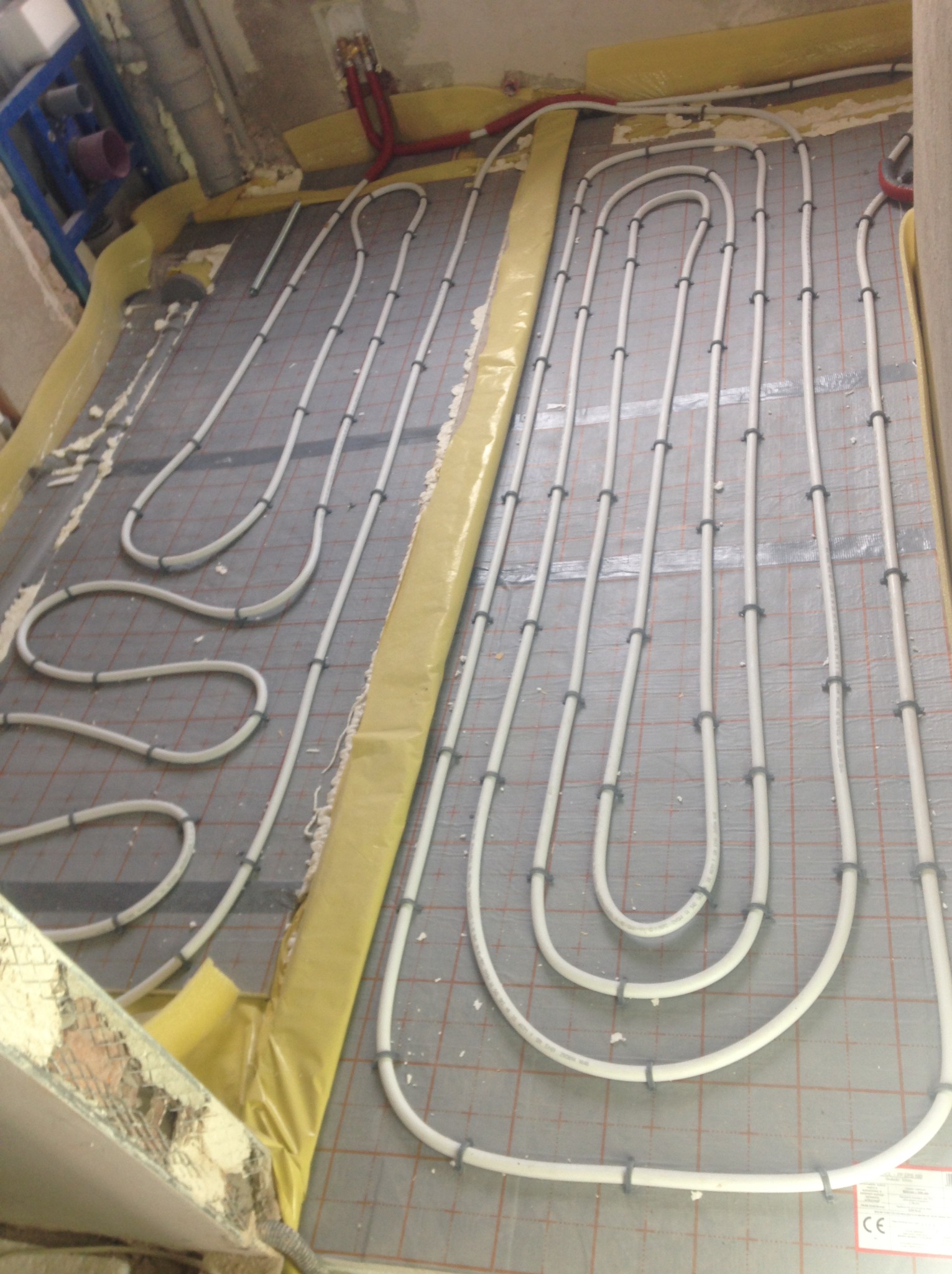 Białe rury ogrzewania podłogowego, zamocowane na szarej macie z siatką, układane w pętle. Żółta folia izolacyjna przy ścianach. Widoczne fragmenty instalacji hydraulicznej.