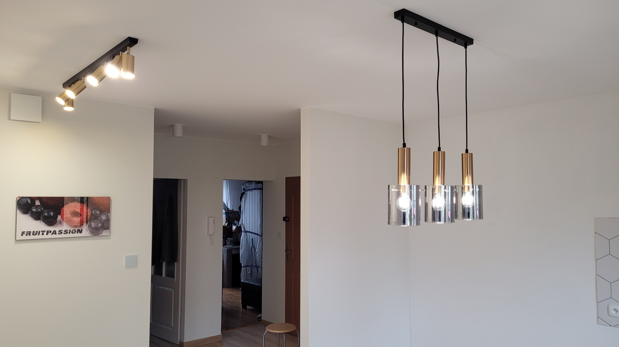 Nowoczesne oświetlenie wnętrza: potrójna lampa wisząca z metalowymi elementami i szklanymi kloszami, oraz reflektory punktowe na suficie, uzupełnione dekoracją ścienną.