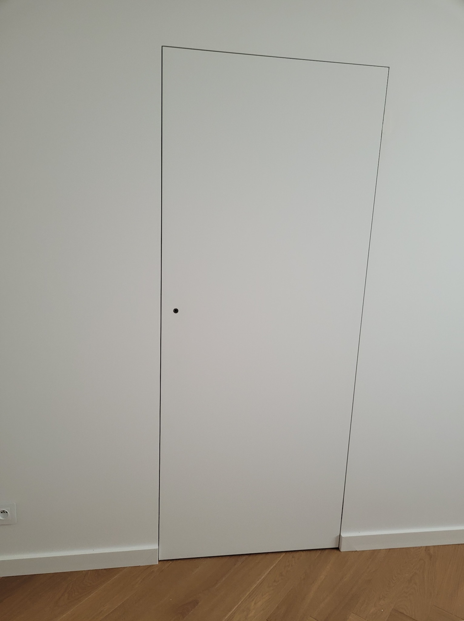 Minimalistyczne, białe drzwi ukryte w ścianie, z drobnym, okrągłym uchwytem, wkomponowane w jasne wnętrze z parkietem. Widoczny fragment gniazdka elektrycznego.