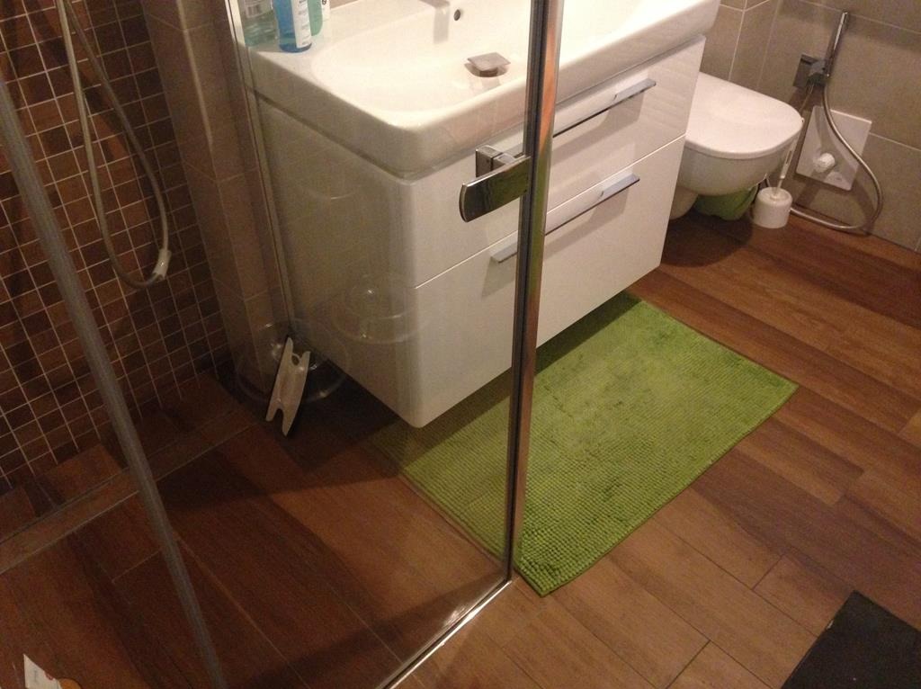 Nowoczesna łazienka w Namysłowie: biała szafka pod umywalką, brązowe płytki imitujące drewno, zielony dywanik i kabina prysznicowa ze szklanymi drzwiami.