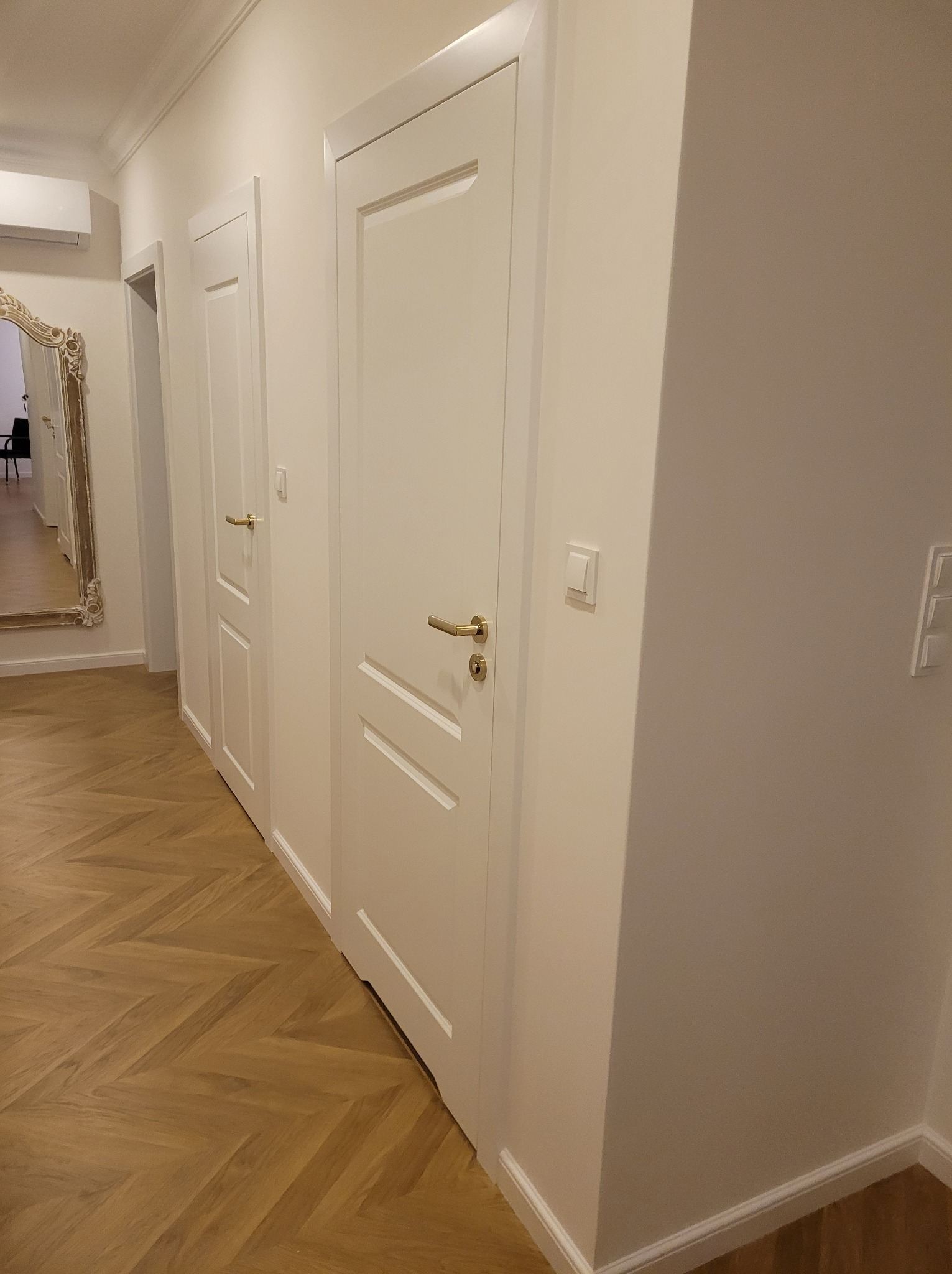 Białe, panelowe drzwi z złotymi klamkami w jasnym korytarzu z parkietem ułożonym w jodełkę. W tle widoczne lustro w ozdobnej ramie i kolejne drzwi.