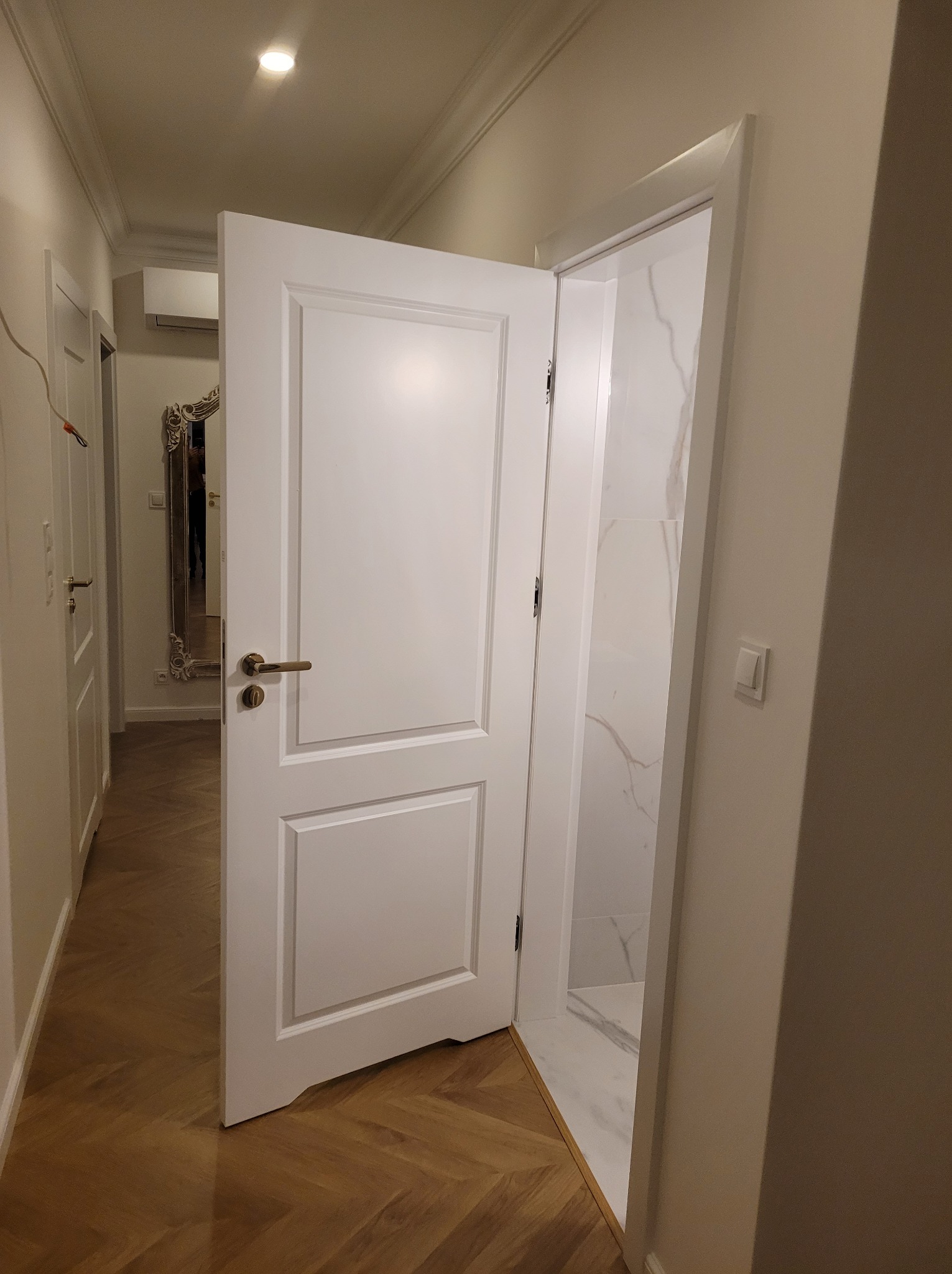 Otwarta, biała, płycinowa  drzwi z mosiężną klamką, prowadząca do łazienki z marmurowym wykończeniem. Parkiet w jodełkę na podłodze. Widok z korytarza.