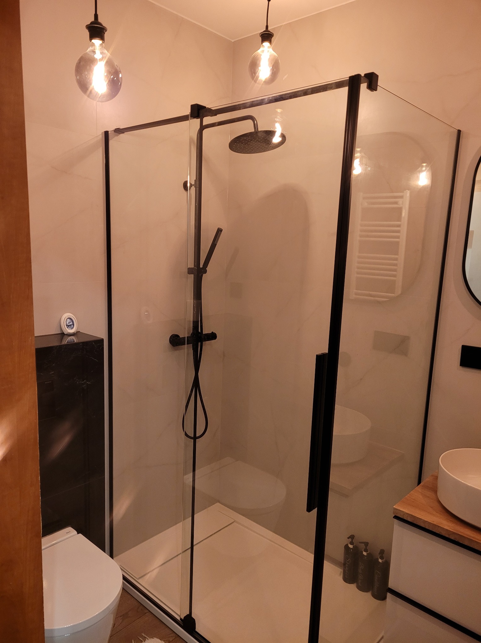 Nowoczesna kabina prysznicowa z czarnymi profilami i deszczownicą w wykończonej łazience z marmurowymi płytkami i wiszącymi lampami w stylu retro.