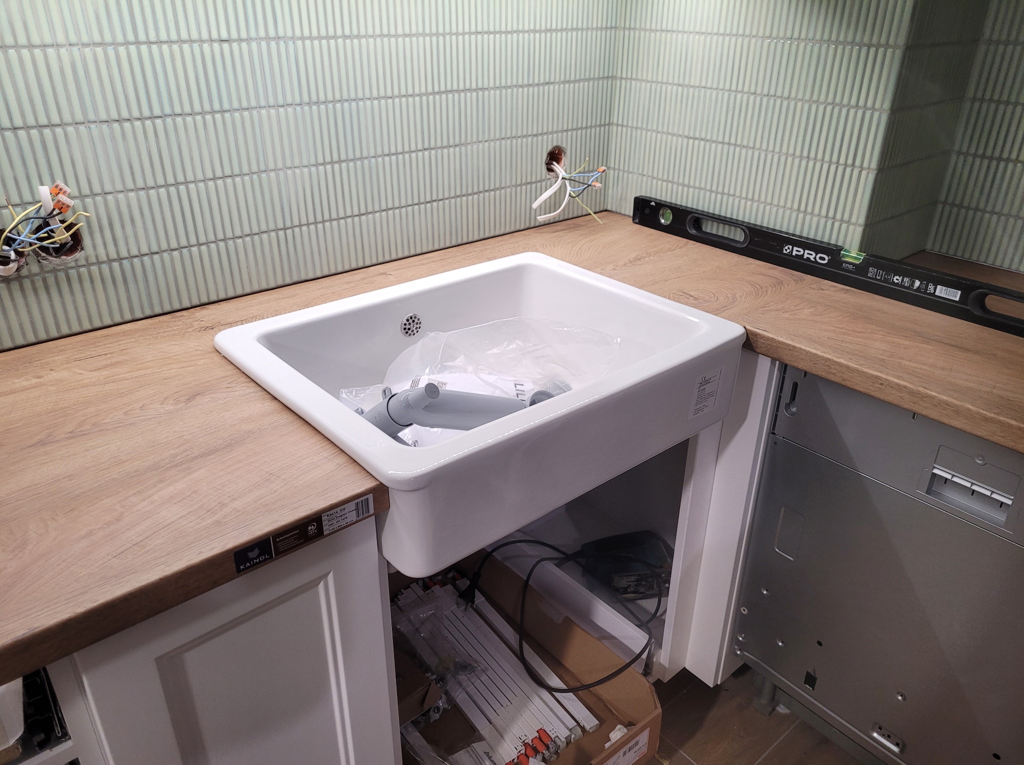 Biała, ceramiczna umywalka wpuszczana w drewniany blat, z widoczną instalacją elektryczną i poziomnicą, na tle ściany wyłożonej podłużnymi, zielonymi płytkami.