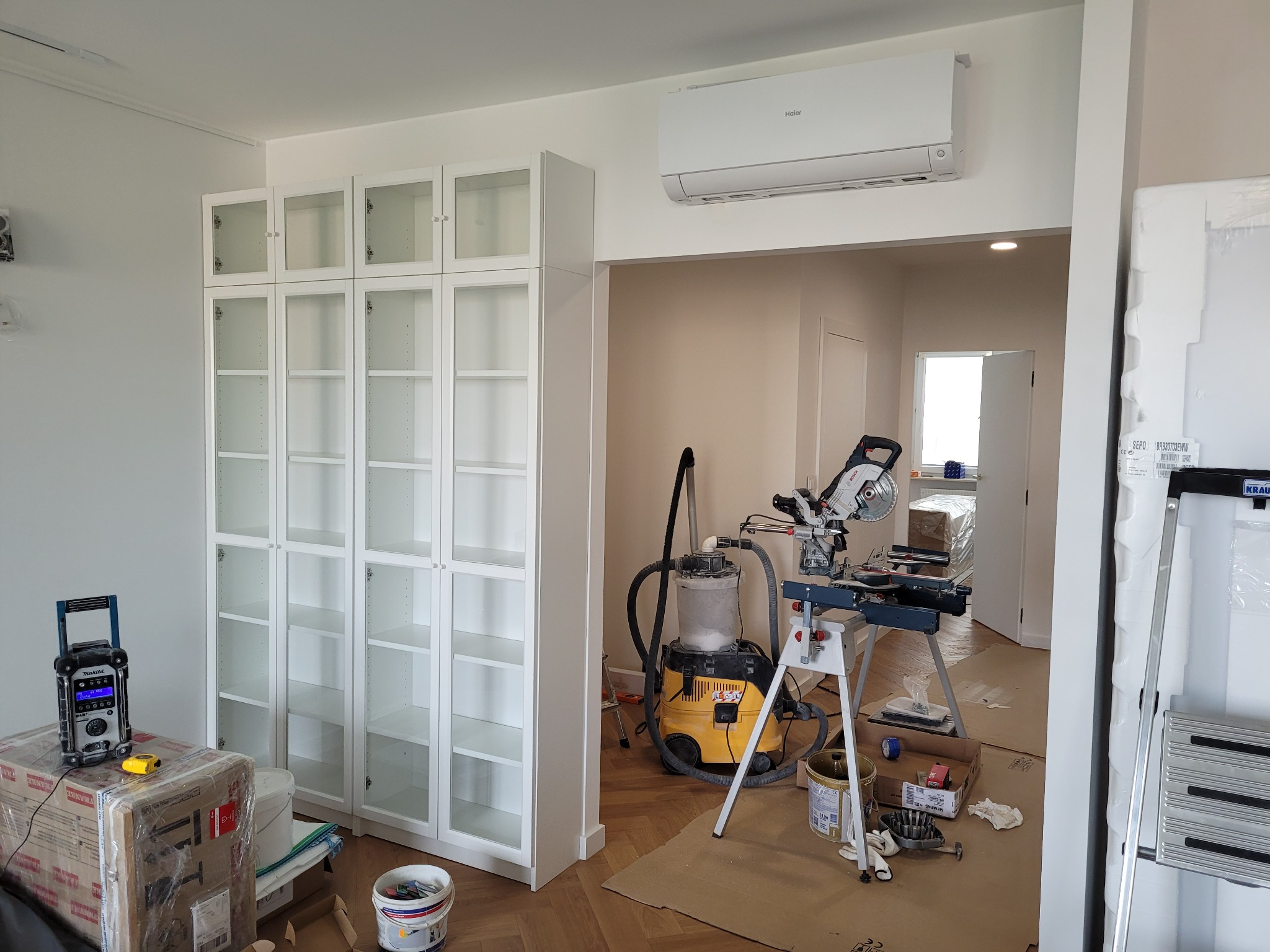 Białe, przeszklone szafki w trakcie montażu, widoczne narzędzia i odkurzacz przemysłowy. Parkiet w jodełkę, klimatyzator na ścianie. Wnętrze w trakcie wykańczania.
