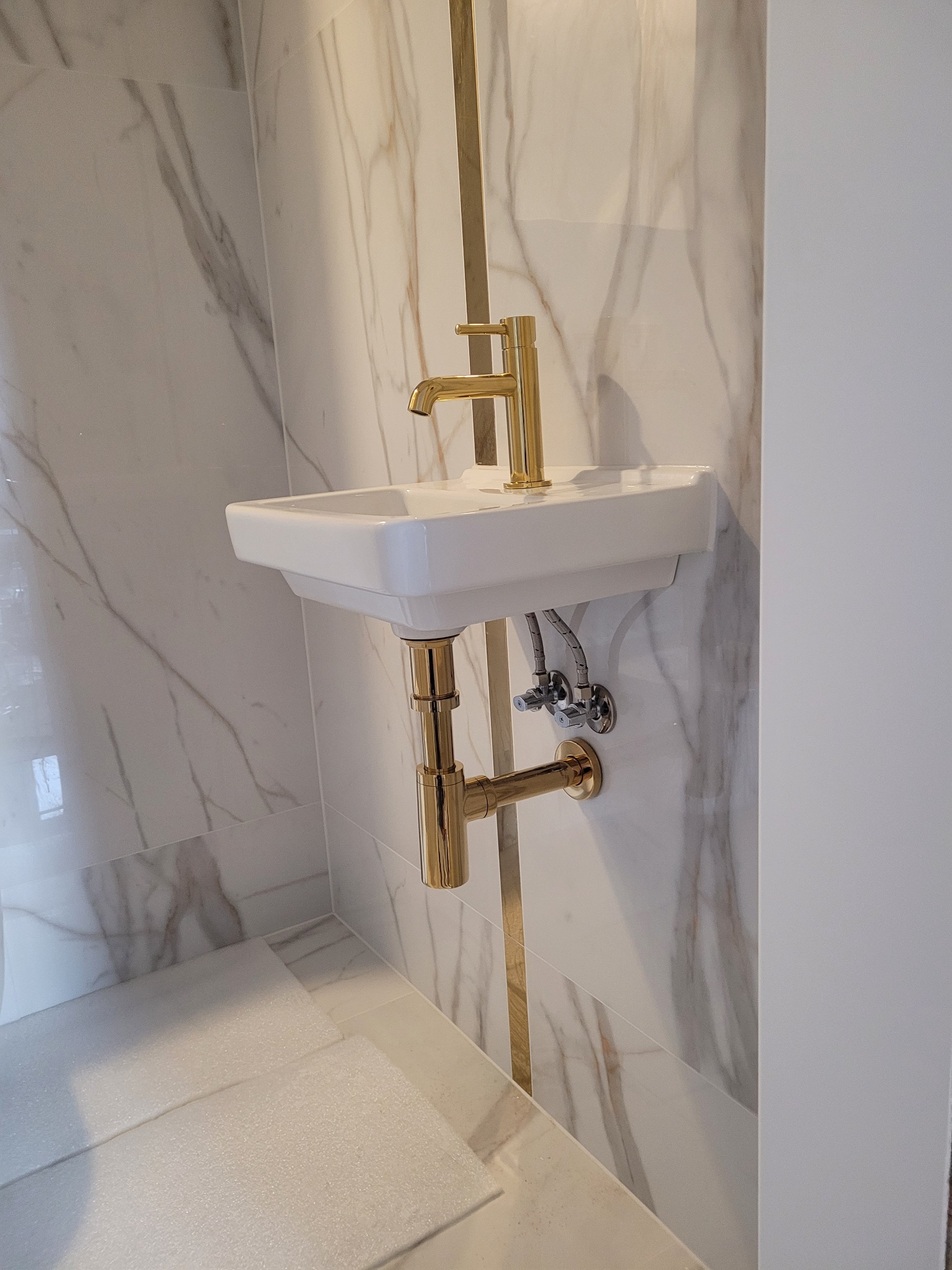 Biała, wisząca umywalka z baterią i syfonem w kolorze złotym, zamontowana na ścianie wyłożonej marmurowymi płytkami. Minimalistyczny design, nowoczesna łazienka.