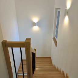 Vintage Look Stolarstwo i Renowacja - Jasne, drewniane schody z czarnymi balustradami i oświetleniem ściennym, widok z góry. Minimalistyczny design, naturalne materiały, nowoczesne lampy.