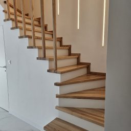 Vintage Look Stolarstwo i Renowacja - Drewniane schody z jasnego drewna z minimalistyczną balustradą i oświetleniem LED na ścianie. Nowoczesny design, białe ściany i marmurowa podłoga.