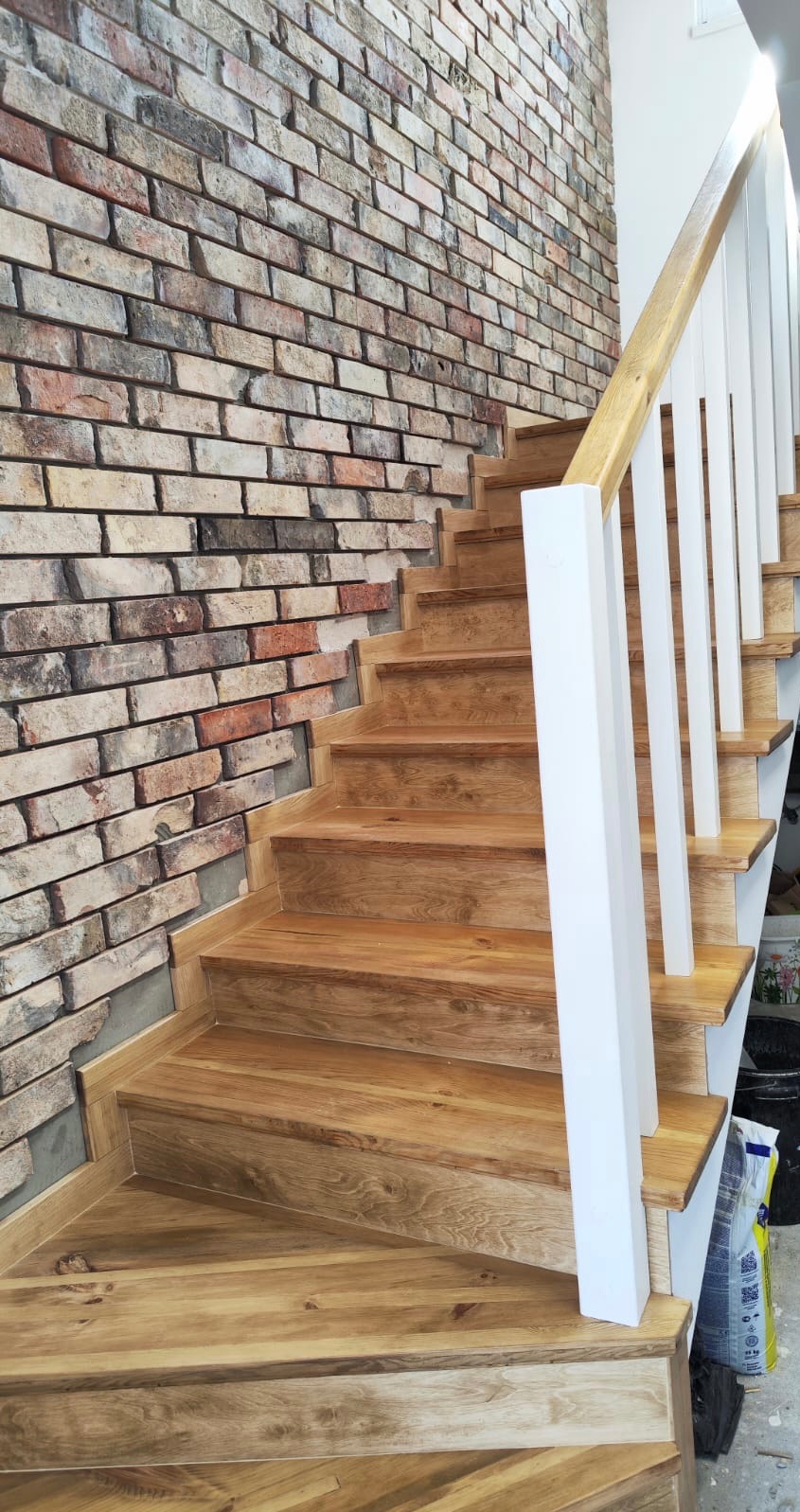 Drewniane schody z białymi balustradami, przylegające do ściany z cegły dekoracyjnej. Widoczne detale wykończenia i naturalna kolorystyka drewna.