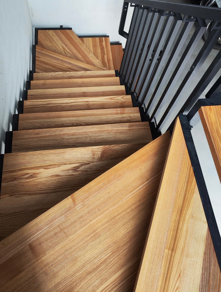 Drewniane schody z metalową balustradą, widok z góry. Stopnie ułożone w jodełkę, jasne drewno z wyraźnym usłojeniem. Nowoczesny design, minimalistyczne wykończenie.